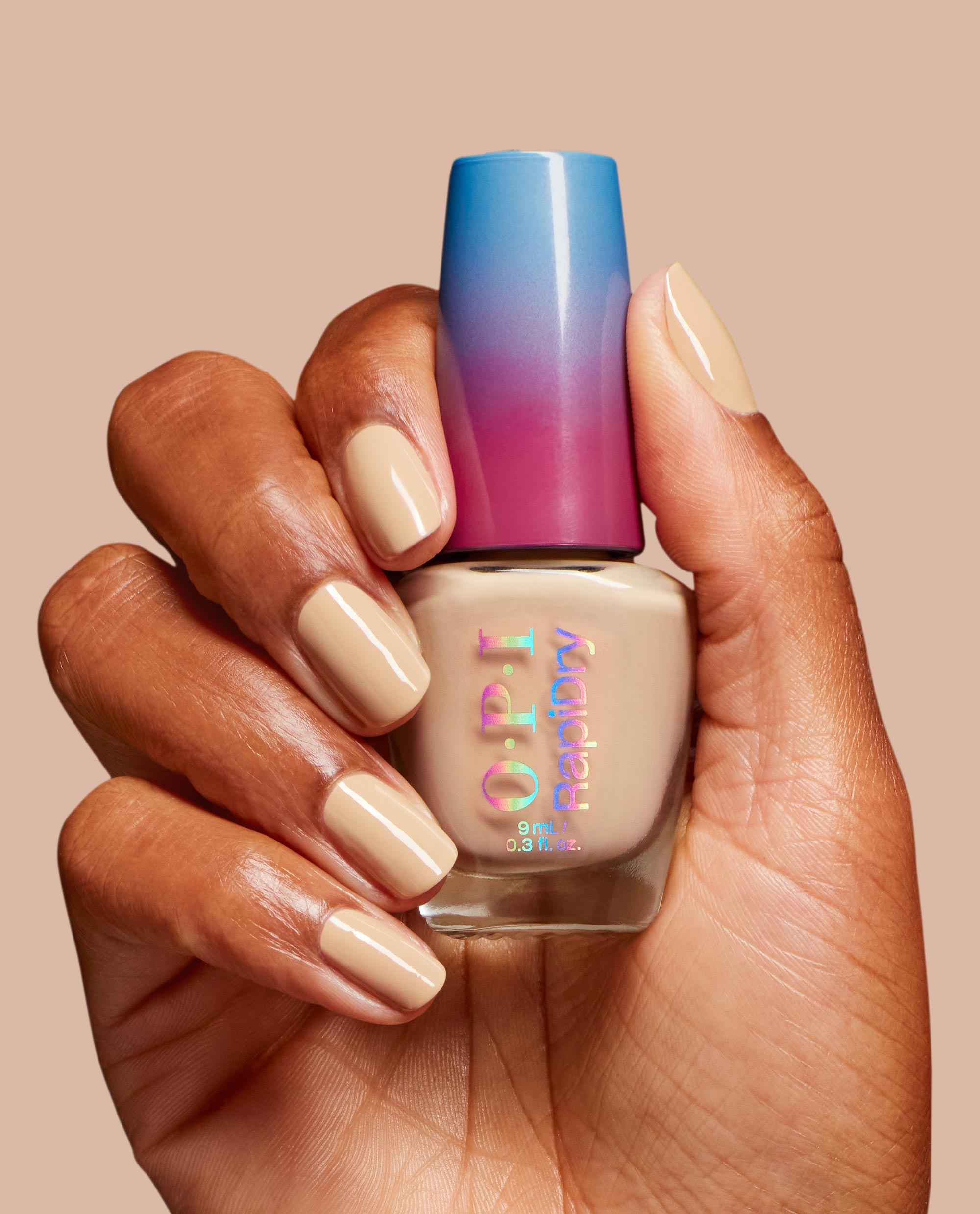 Brown hand holds OPI RapiDry nail polish in Can't Hardly Wait, a trending glossy beige nude shade matching manicured nails, with a blue-to-magenta cap on a warm neutral background.