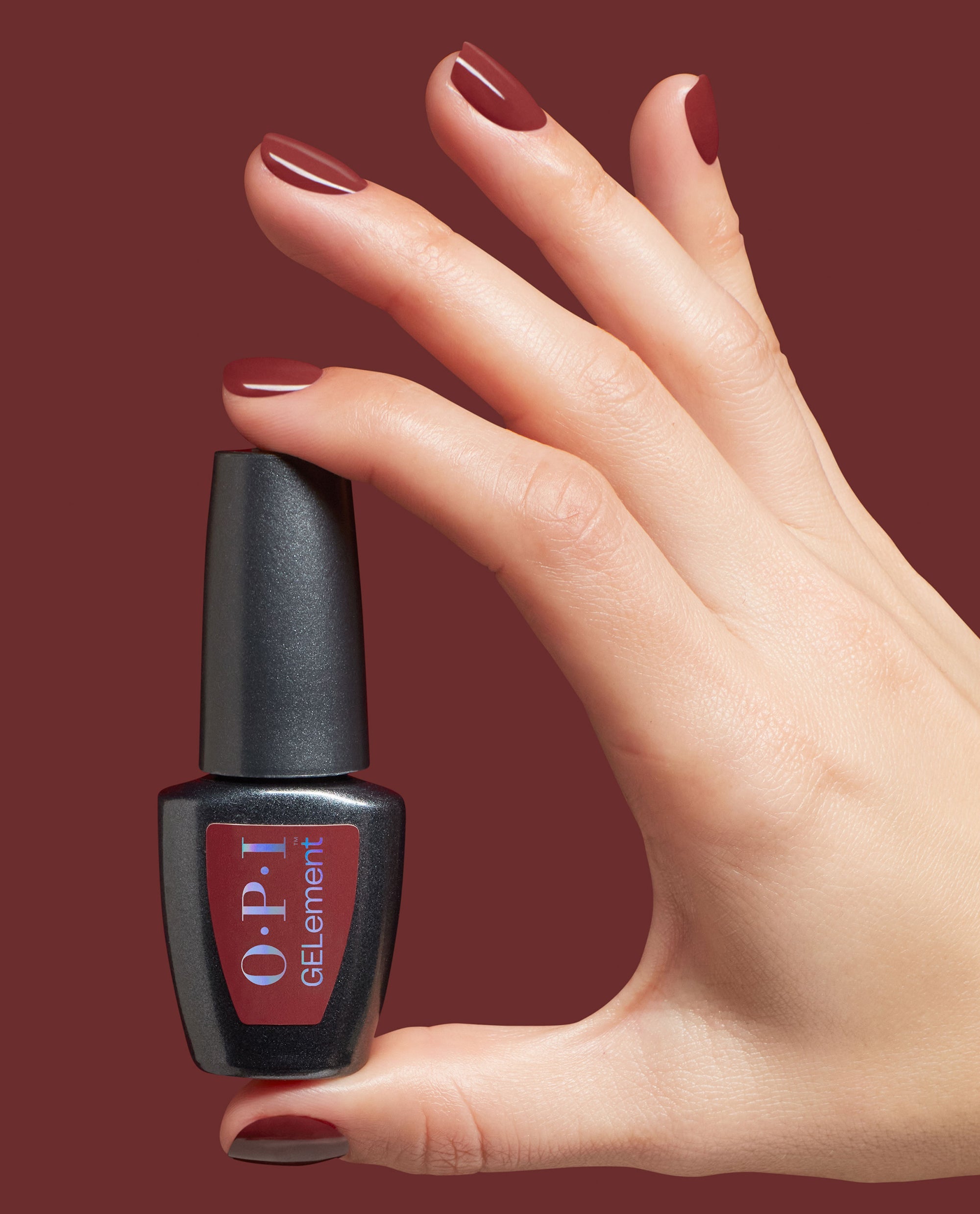 Hand with glossy burgundy nails holds OPI GelColor Como Se Dice Gel-ous? at-home gel nail polish, a rich red-brown shade for fall nail colors, against a matching maroon background.