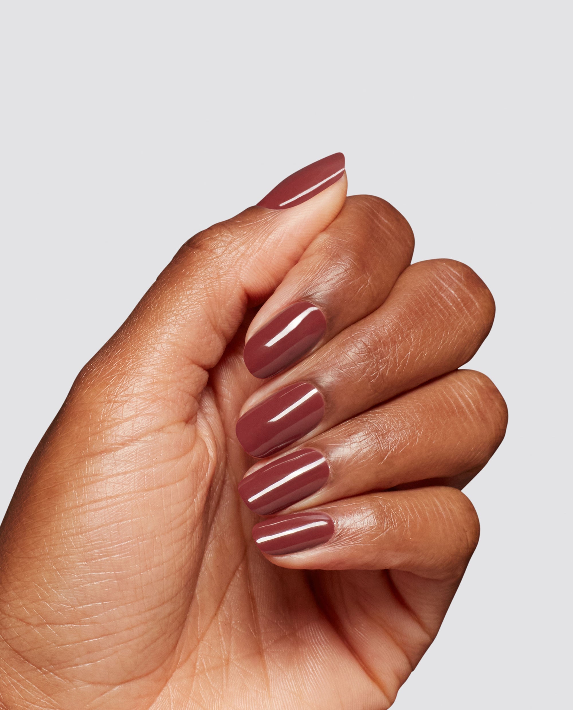 Hand modeling glossy mauve-brown Como Se Dice Gel-ous? at-home gel nail polish by OPI, showing rounded fall nail colors on a clean light gray background.