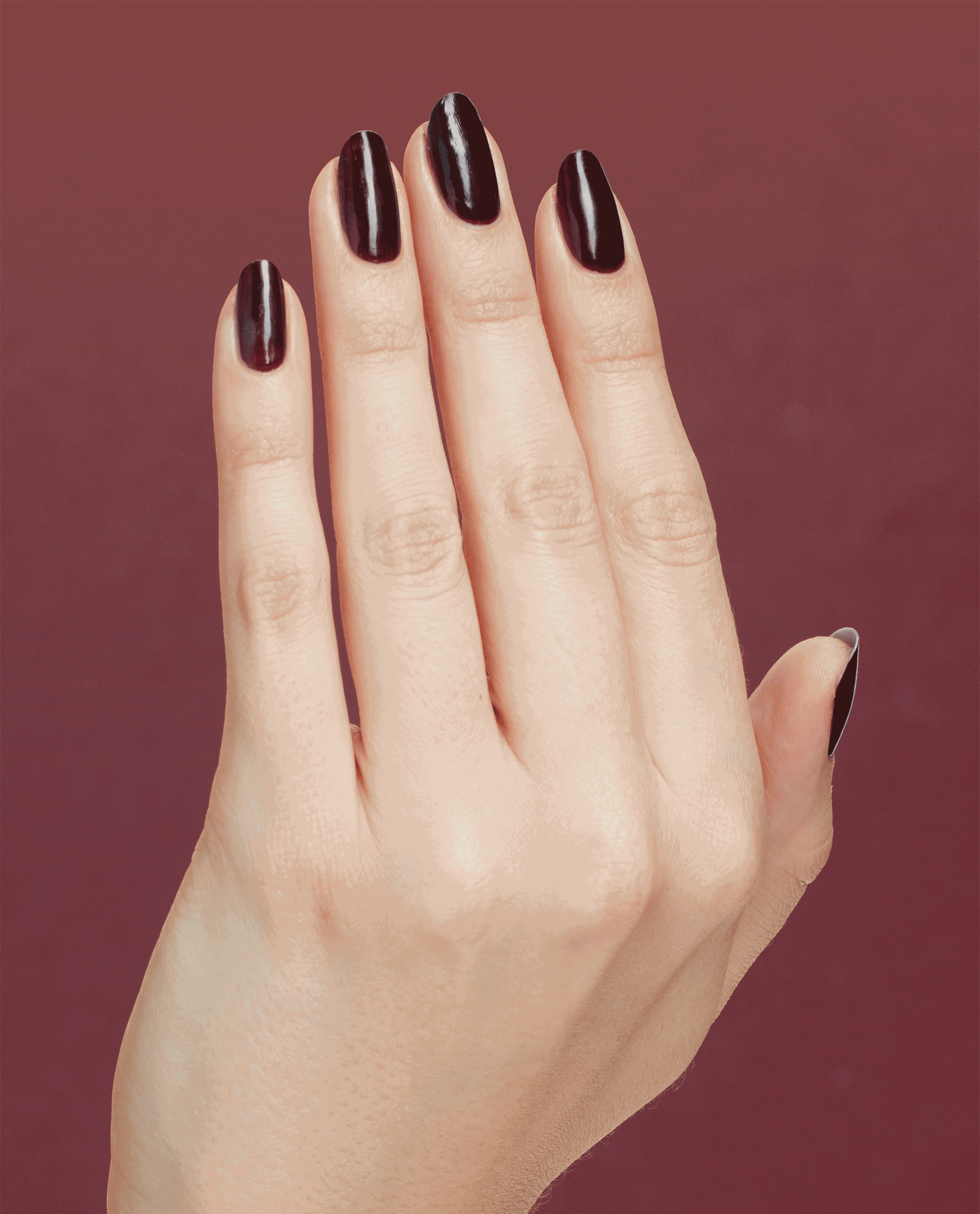 Hand with long oval nails painted in Complimentary Wine, a deep burgundy OPI nail polish nail lacquer, flexing to show glossy, even coverage against a coordinating wine-red background.