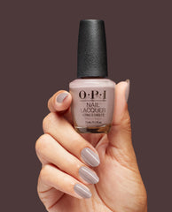 Hand with glossy taupe-beige manicured nails holding an OPI nail polish bottle, shade Crimp and Proper nail lacquer, against a solid brown background, showcasing the neutral nude color and smooth finish for US shoppers.