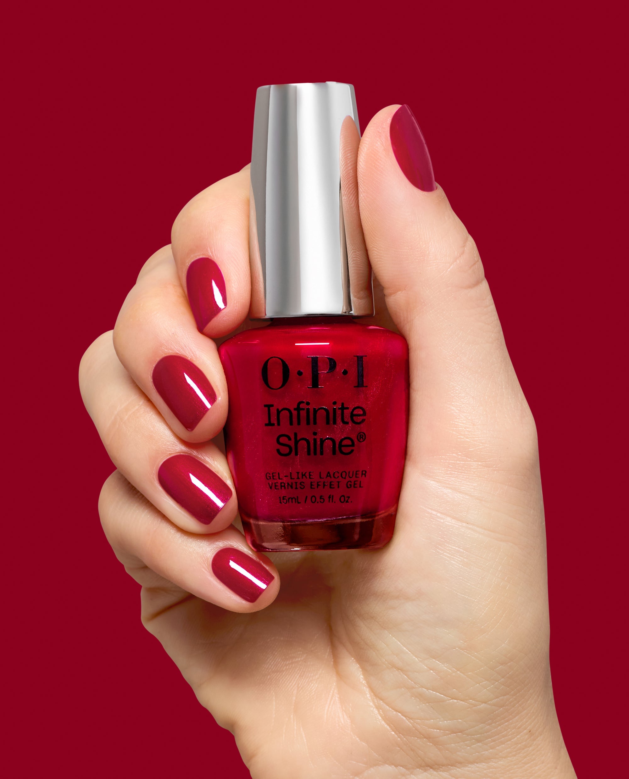 Hand with glossy deep berry red nails holds OPI Infinite Shine Deer Valley Spice gel-like lacquer, a rich shade for fall nail colors, against a matching red background.