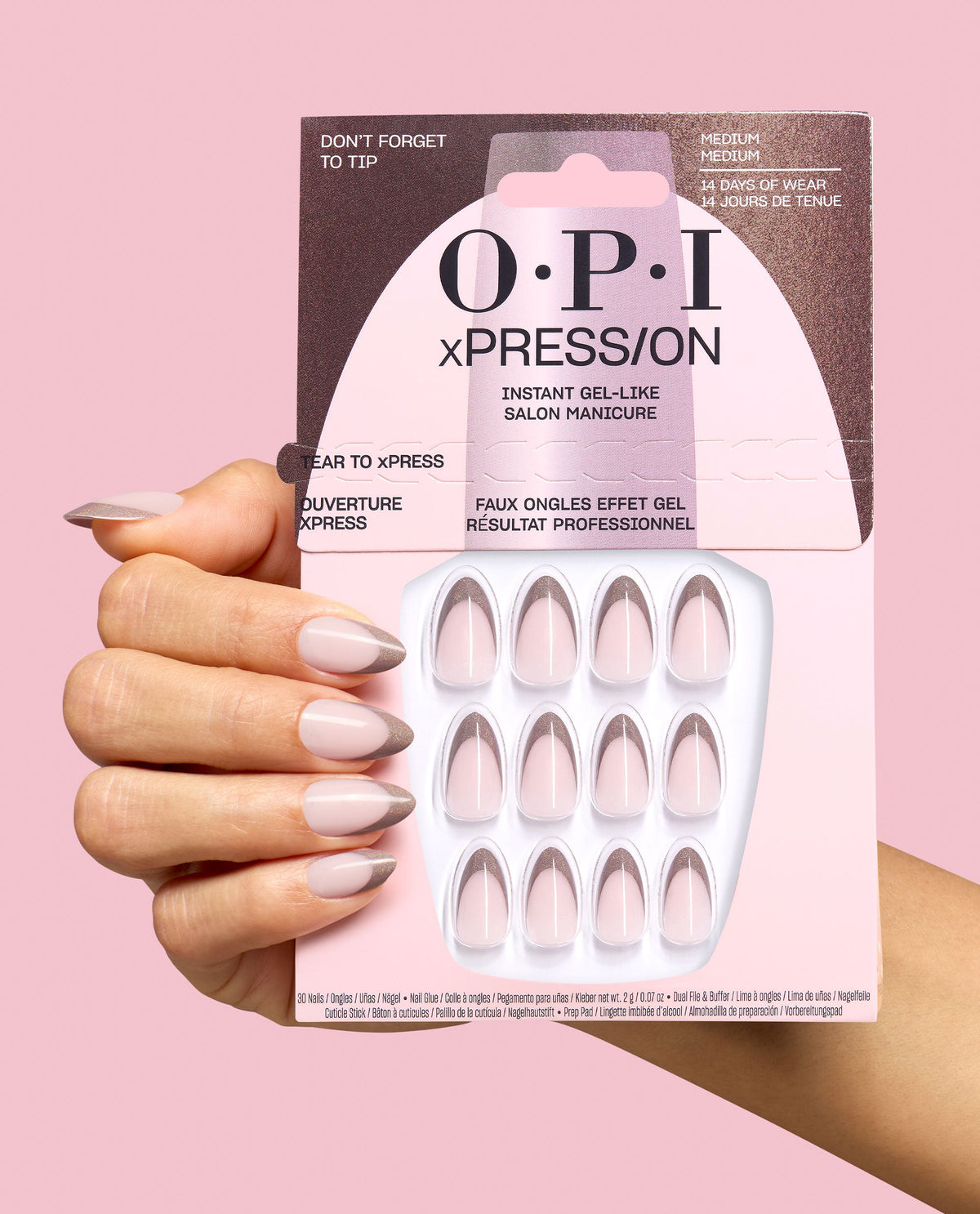 Hand holds OPI xPRESS/ON Don’t Forget to Tip press-on nails in pale pink with metallic taupe French tips, shown in retail packaging against a pink background for chic fall nail colors.
