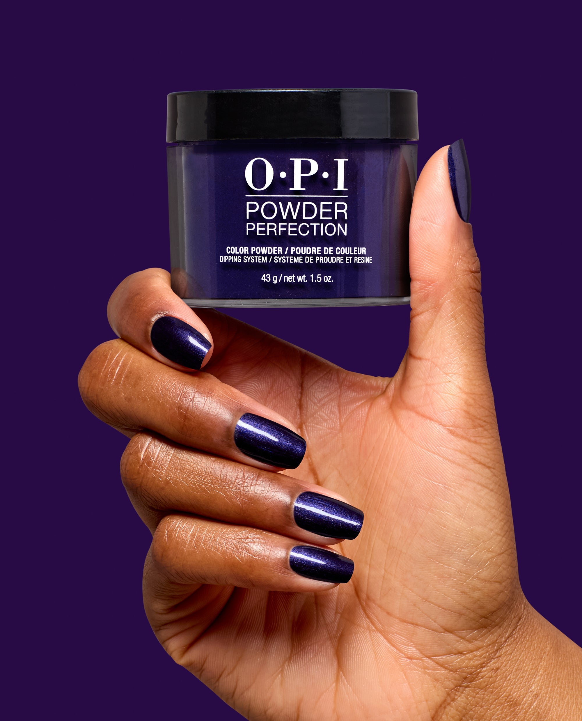 Hand with glossy deep purple nails holds OPI Powder Perfection Don’t Over Ink It dipping powder, a rich shade for fall nail colors, against a solid purple background.