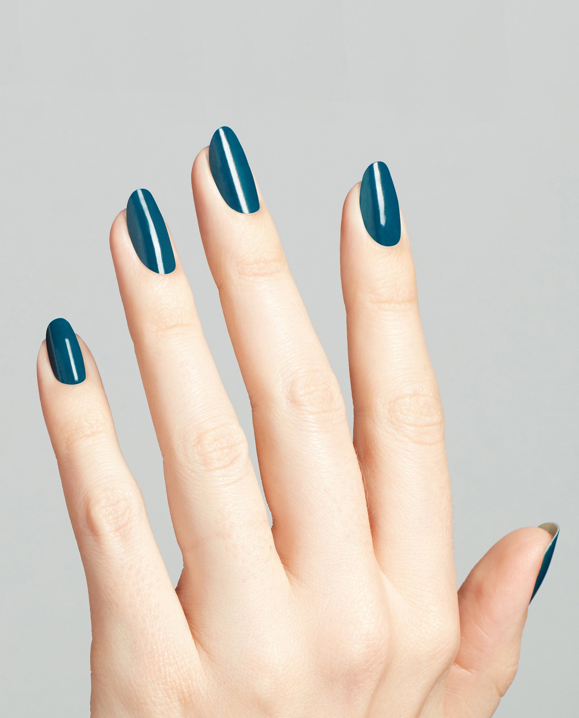 Hand with long oval nails painted in Drama at La Scala, a glossy deep teal OPI nail polish, shown against a clean light gray background to highlight the rich color and smooth nail lacquer finish.