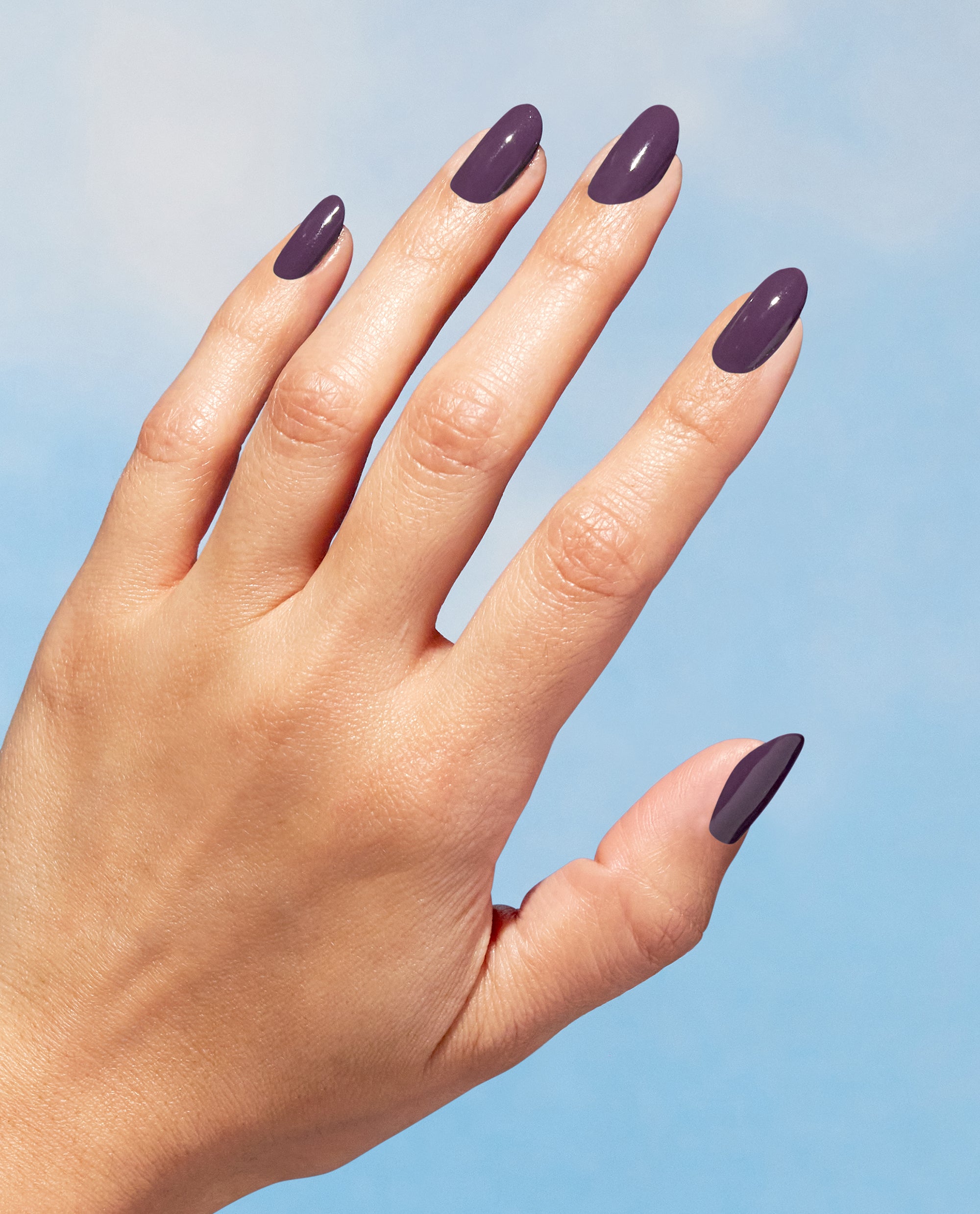 Hand modeling glossy deep plum Eco-Maniac OPI Nature Strong Vegan Nail Lacquer on almond-shaped nails against a blue background, a rich pick for fall nail colors.
