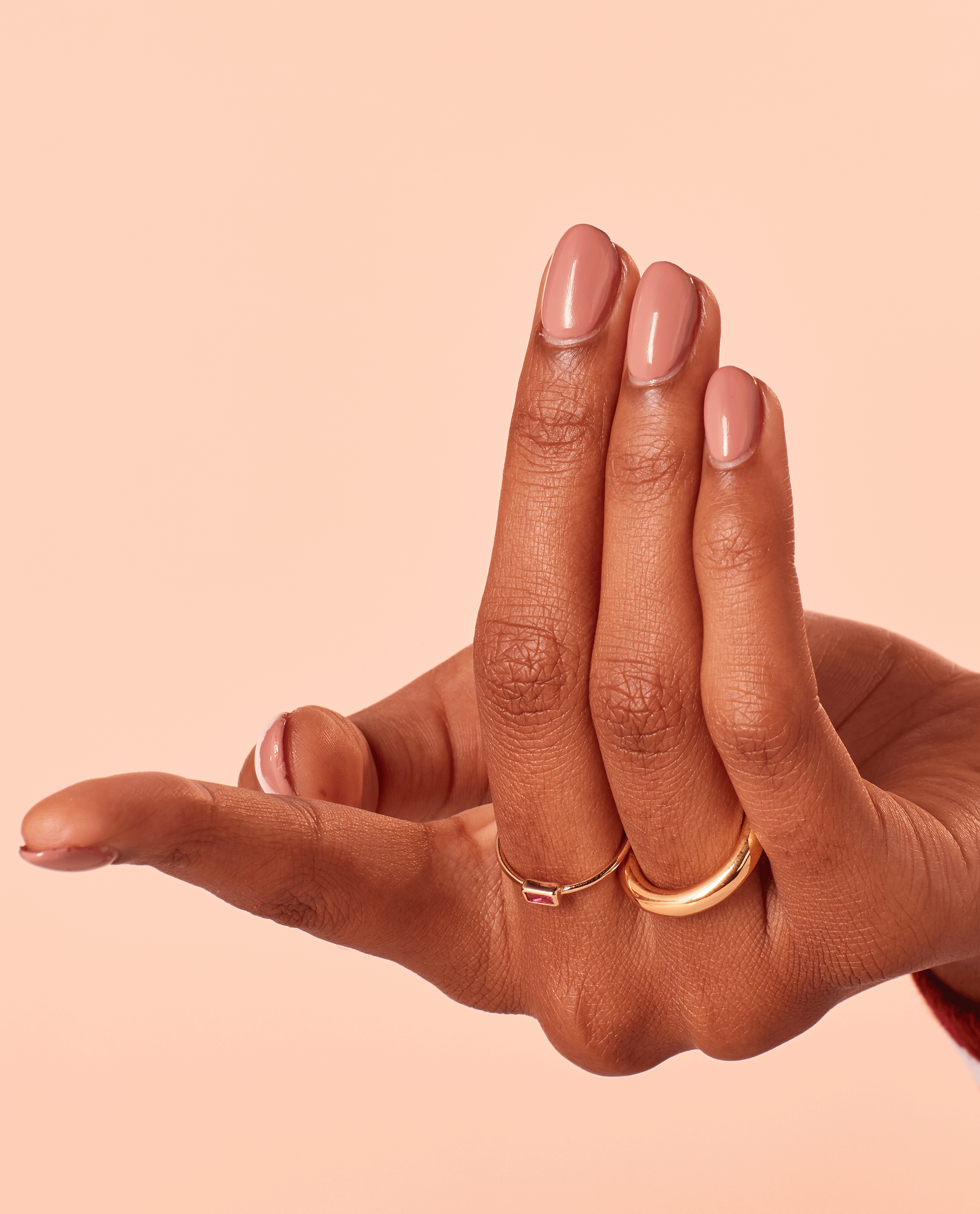 Hand with short, oval nails painted in Edinburgh-er & Tatties OPI nail polish, a glossy pinky‑nude nail lacquer, fingers slightly bent to showcase the smooth finish against a peach background with delicate gold rings.