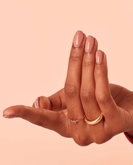 Hand with short, oval nails painted in Edinburgh-er & Tatties OPI nail polish, a glossy pinky‑nude nail lacquer, fingers slightly bent to showcase the smooth finish against a peach background with delicate gold rings.