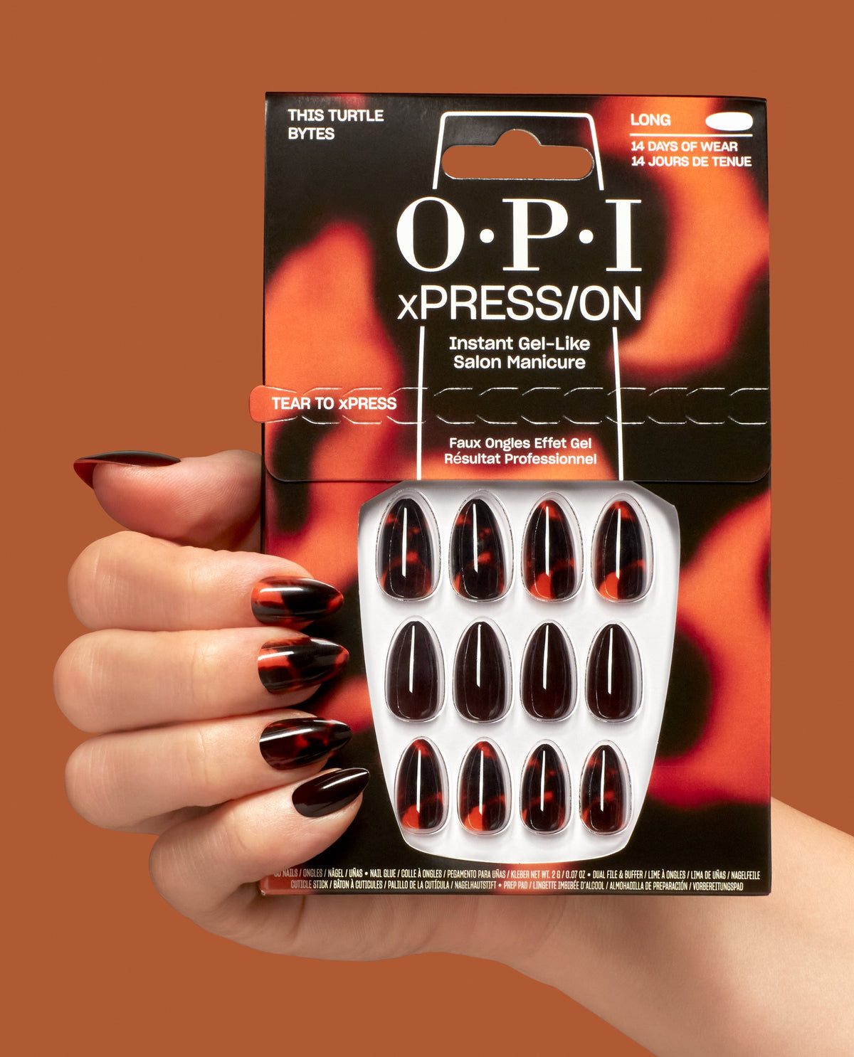 A hand wearing OPI xPRESS/ON This Turtle Bytes press-on nails holds the pack, showing long gel-like tortoiseshell black and burnt orange fall nail colors on a warm brown background.
