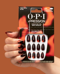 A hand wearing OPI xPRESS/ON This Turtle Bytes press-on nails holds the pack, showing long gel-like tortoiseshell black and burnt orange fall nail colors on a warm brown background.