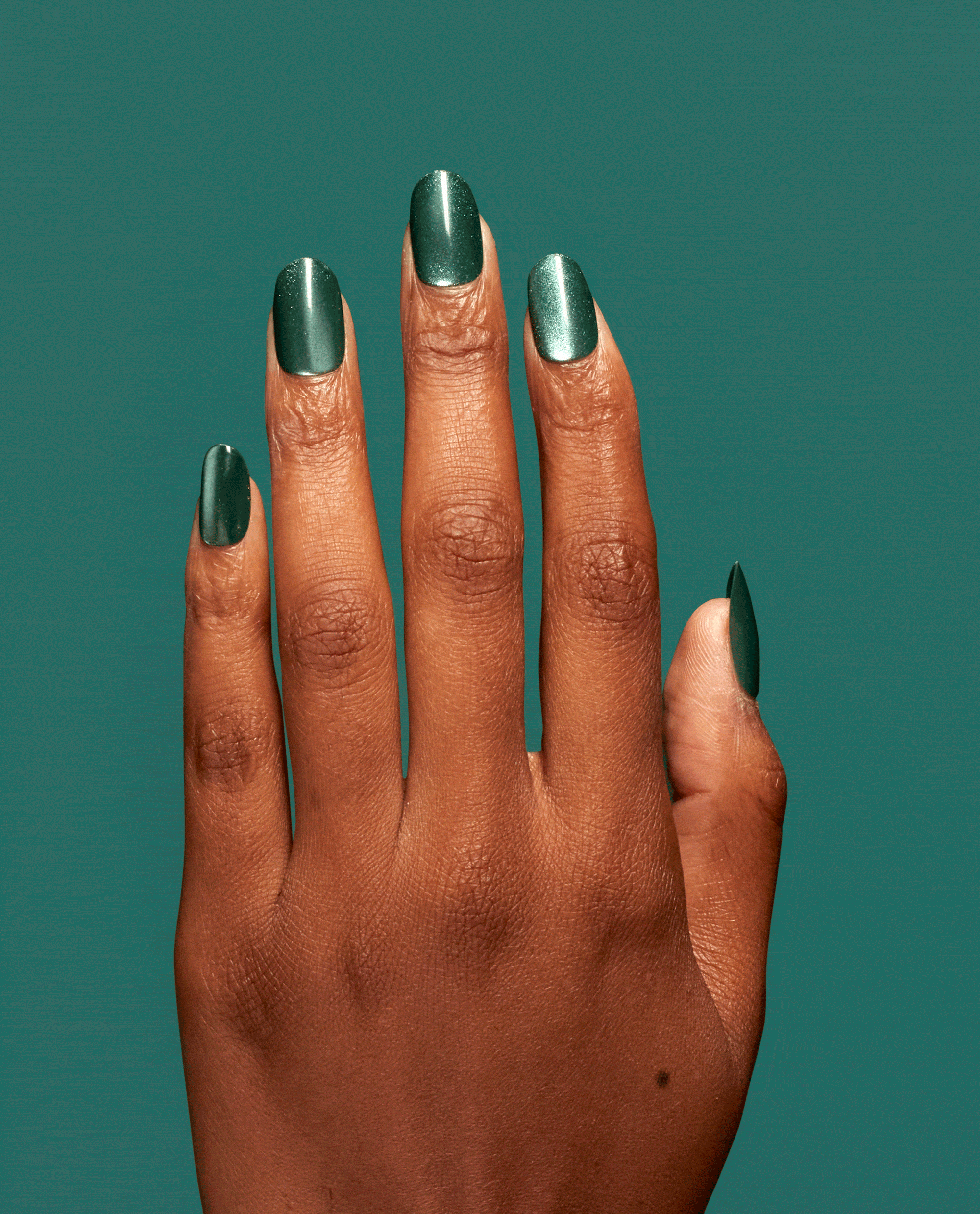 Hand modeling OPI Feelin' Capricorn-y xPRESS/ON press-on nails in a deep emerald green shimmer shade with glossy almond tips against a teal background.