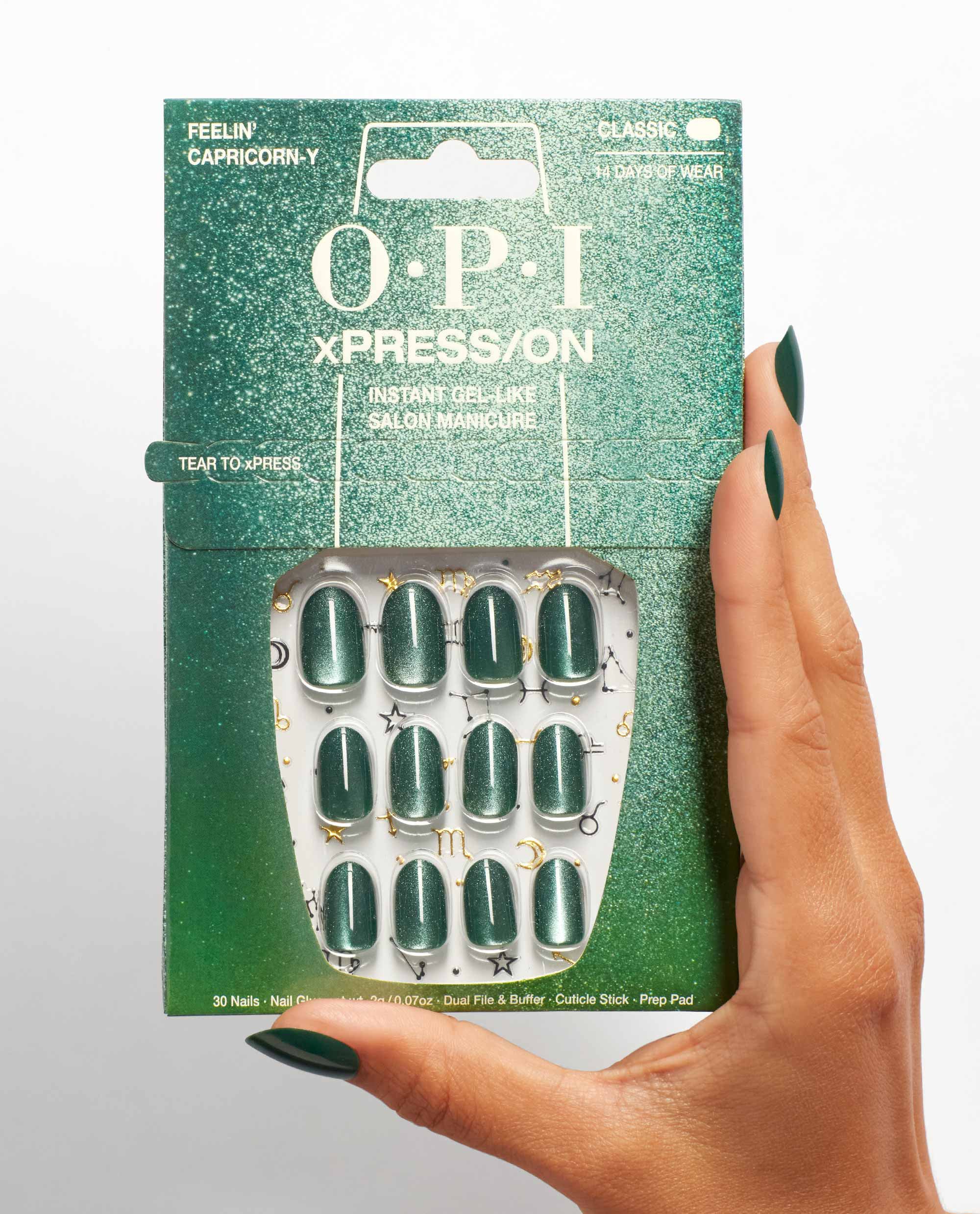 Hand with dark green almond nails holds OPI xPRESS/ON Feelin’ Capricorn-y press-on nails, showing emerald shimmer nails in glittery green packaging with zodiac accents.