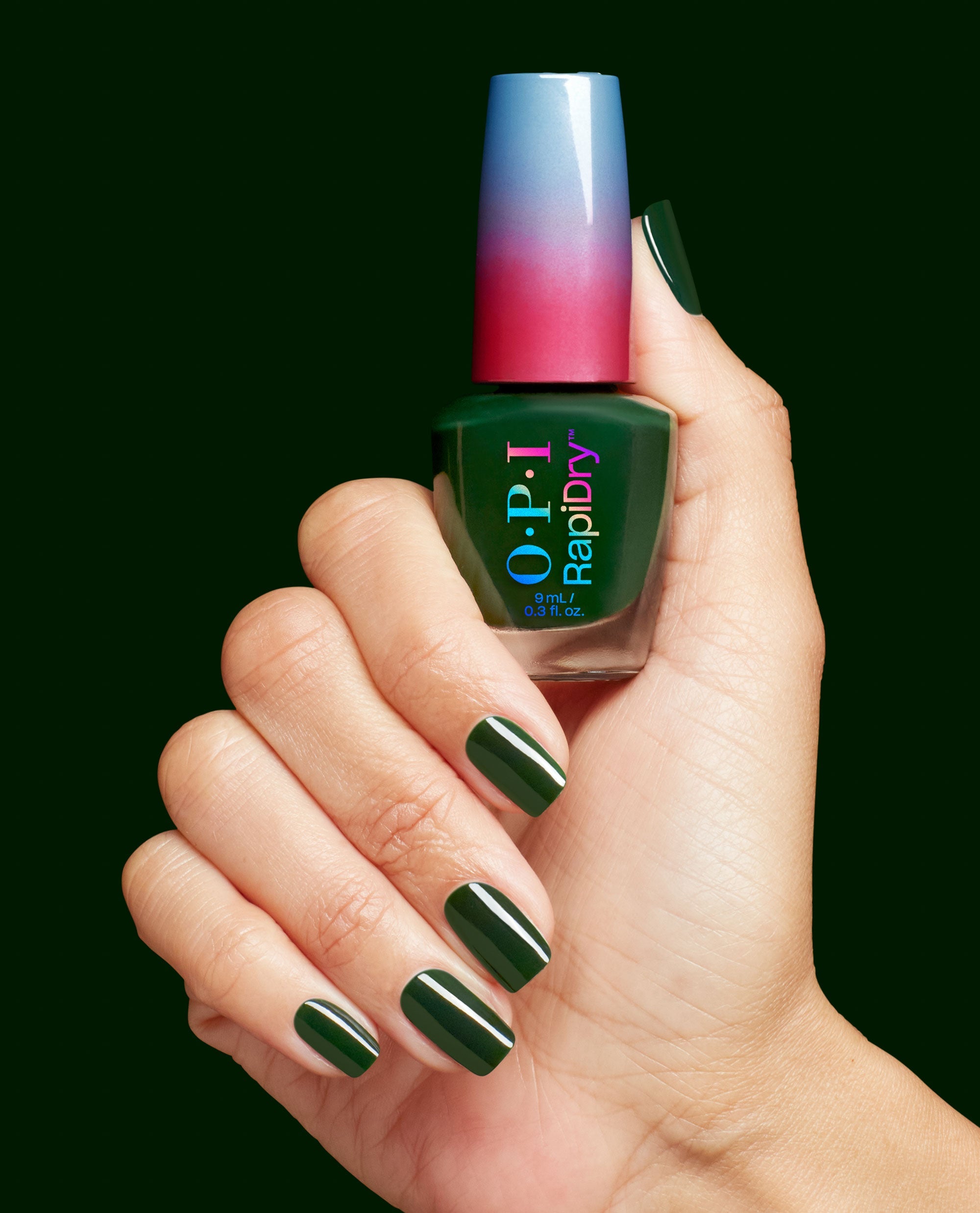 Hand with glossy deep forest green nails holds an OPI RapiDry nail lacquer bottle in Fir Surely Fast, featuring a blue-to-red ombré cap against a dark green background.