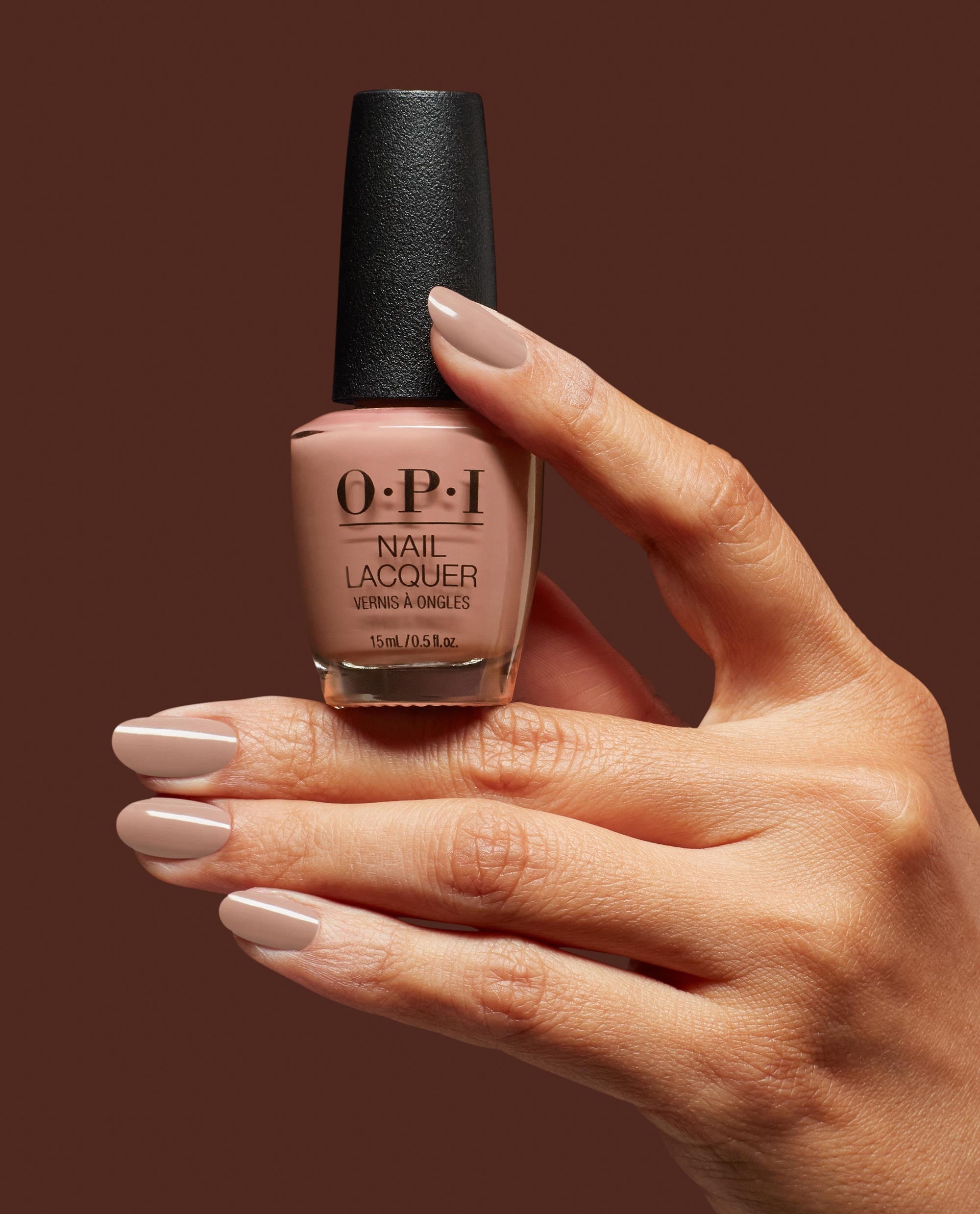 Hand with manicured nails painted in OPI nail polish shade Flannel Aerobics holding the matching OPI Nail Lacquer bottle against a rich brown background, showcasing the creamy neutral beige-pink color and glossy finish.