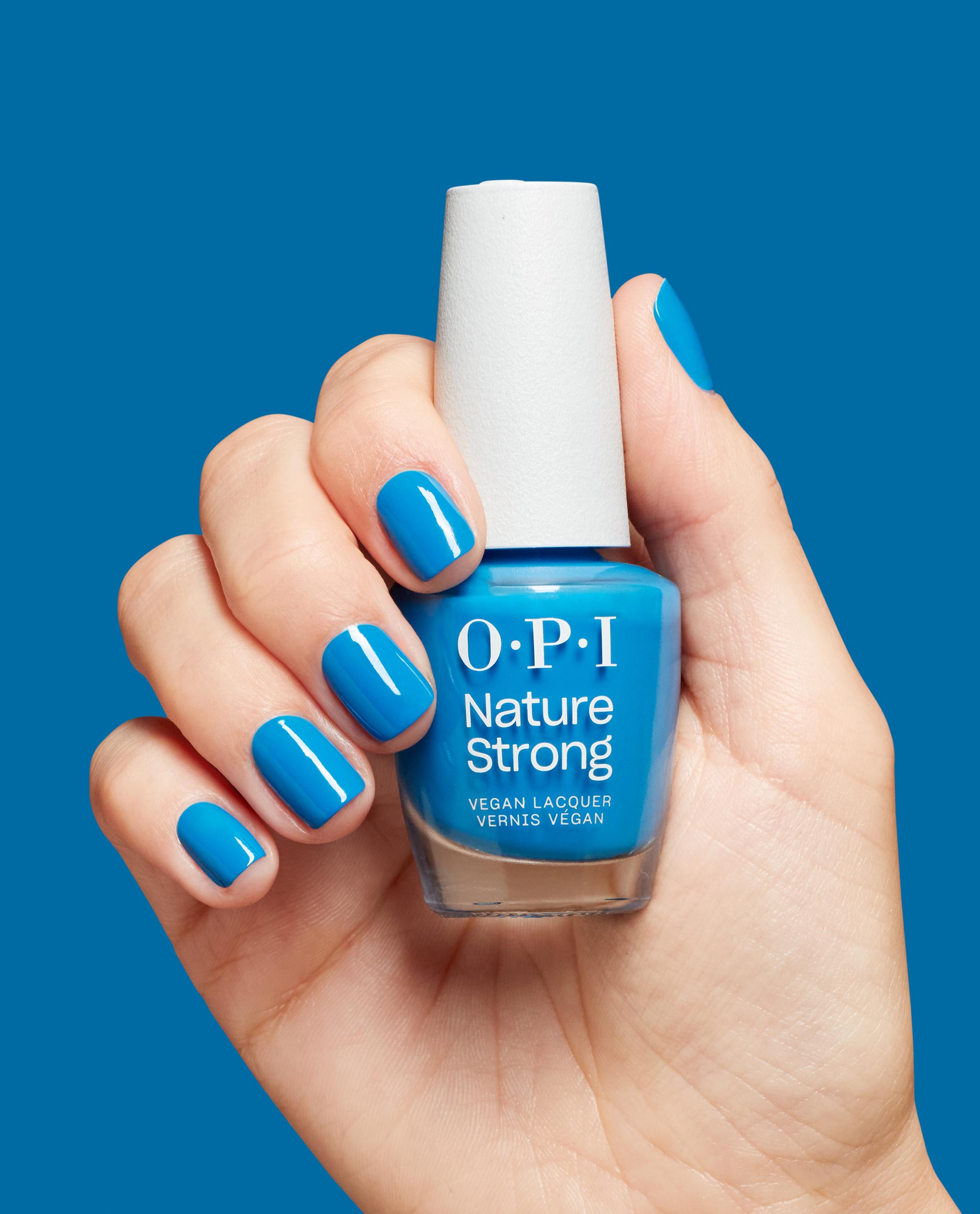 Hand holding OPI Nature Strong Vegan Nail Lacquer in Fresh as a Blue Daisy, with glossy bright blue manicured nails against a matching blue background.