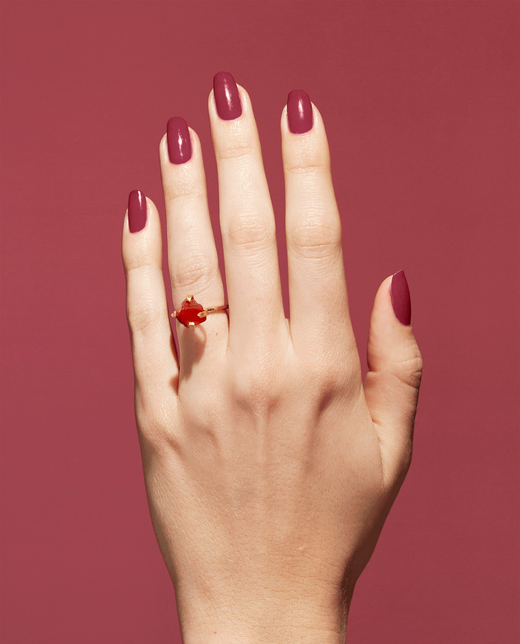 Hand displays glossy garnet-red nails polished with OPI Nature Strong Vegan Nail Lacquer in Give a Garnet, paired with a red gemstone ring on a matching red background for fall nail colors.