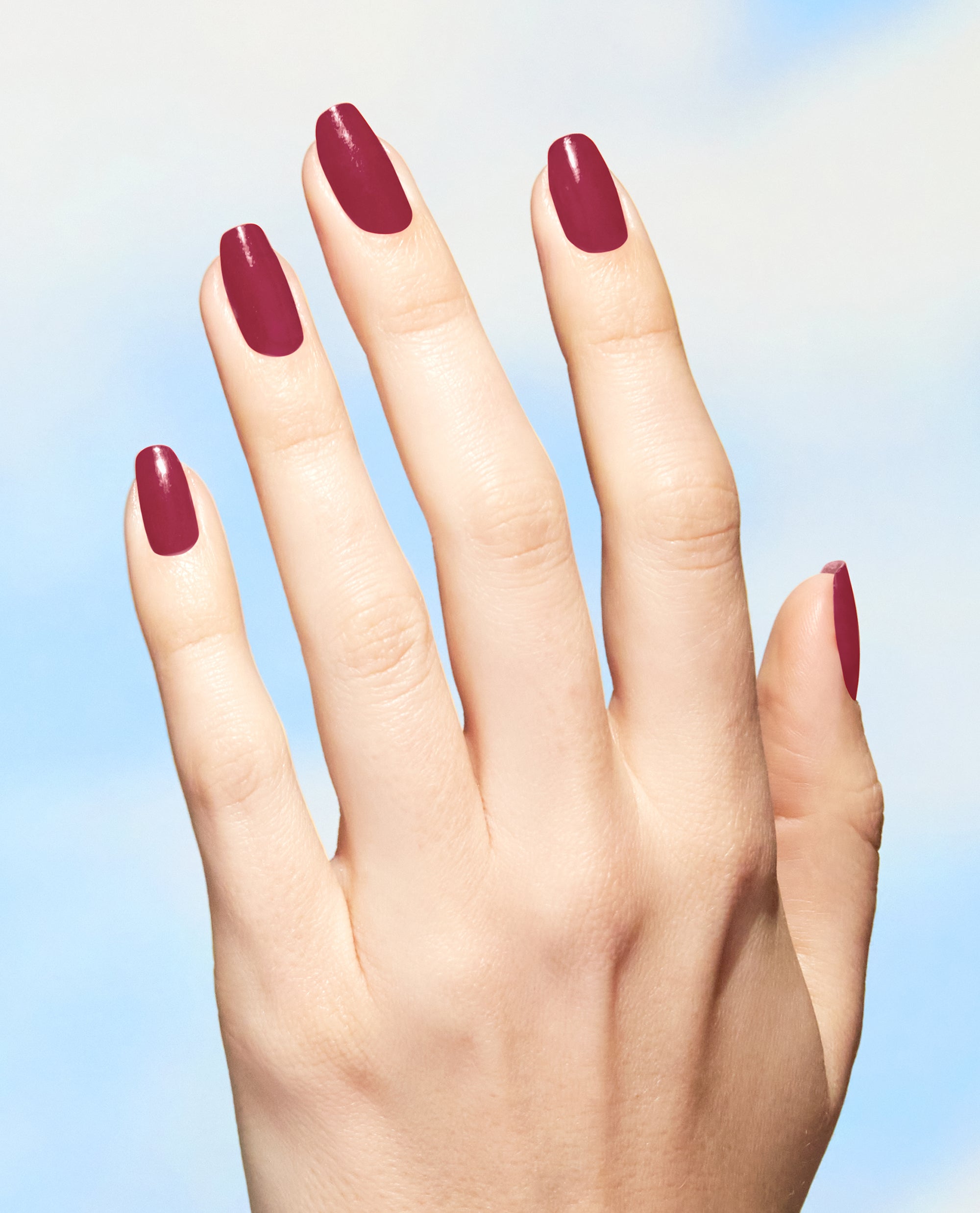 Hand displays glossy garnet-red nails painted with OPI Give a Garnet Nature Strong Vegan Nail Lacquer, a rich choice for fall nail colors against a soft blue background.