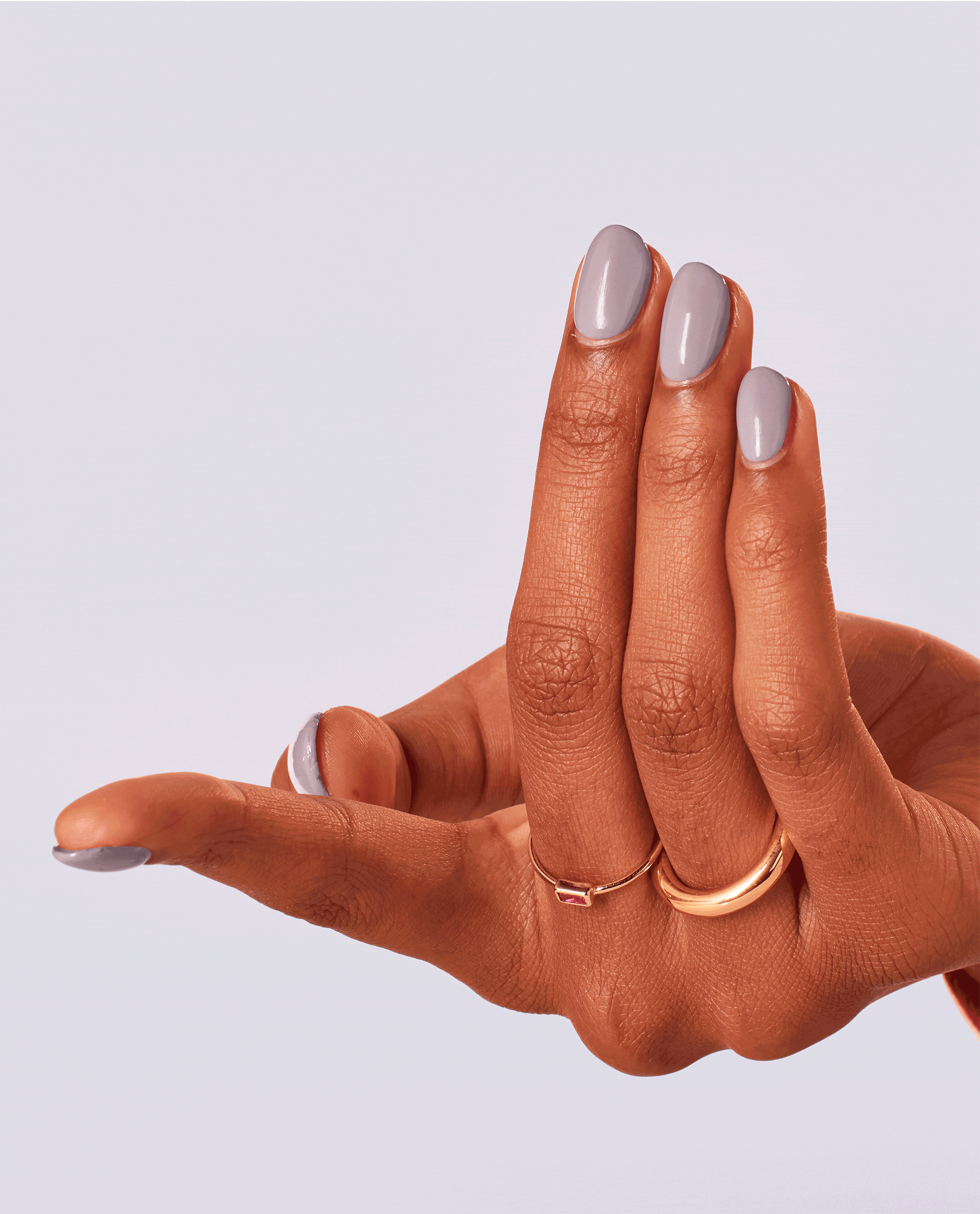 Hand with medium skin tone shows glossy pastel lavender Graffiti Sweetie OPI nail polish manicure, nails shaped oval and paired with delicate gold rings, against a clean light purple background showcasing the nail lacquer color and shine.
