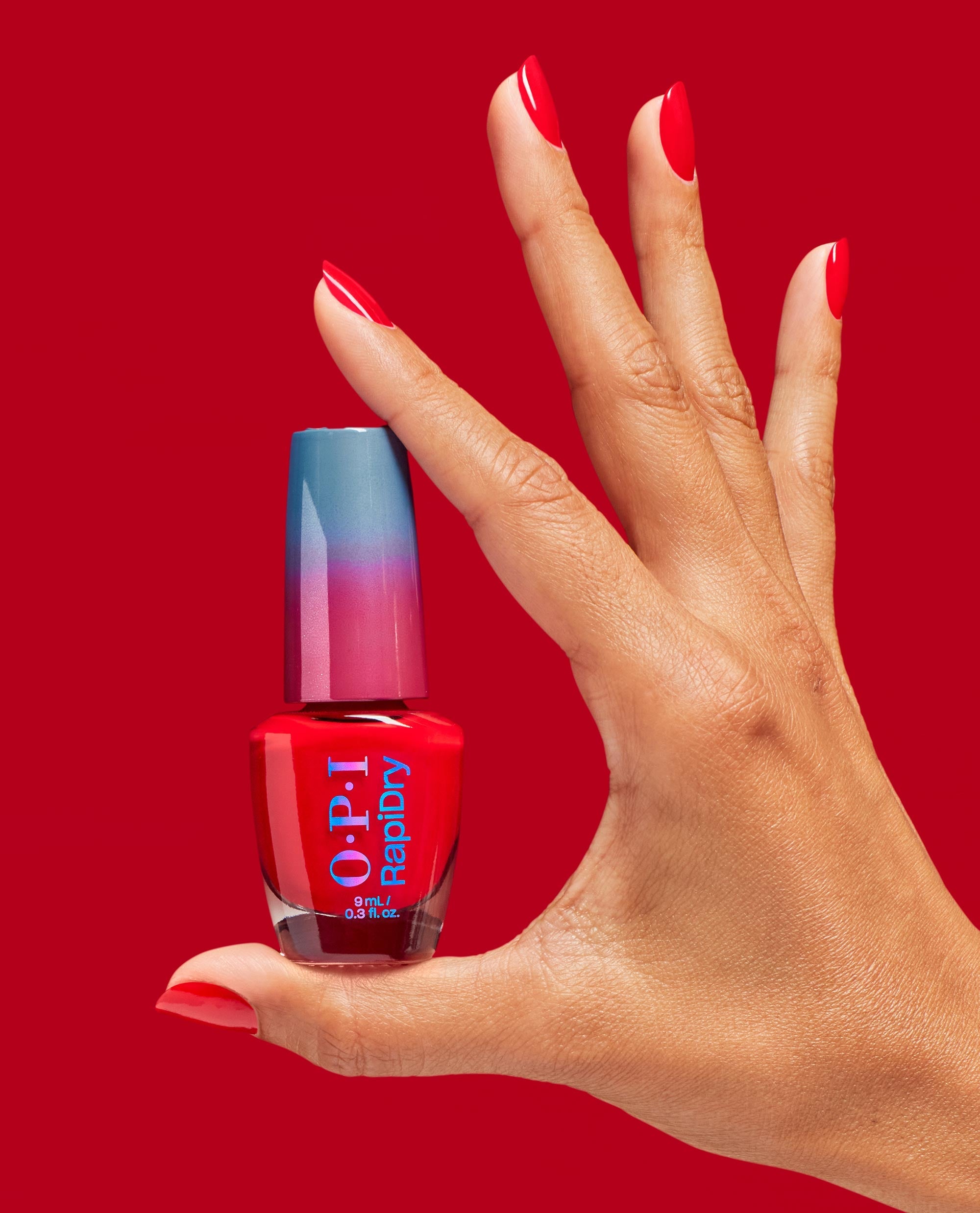 Hand with glossy red almond nails holds OPI RapiDry nail polish in Haul Sass, a vivid red shade for fall nail colors, against a matching red background.