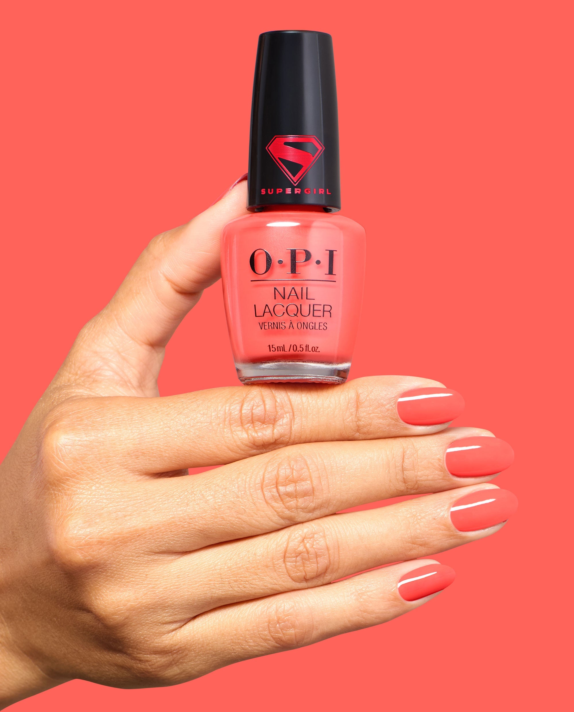 Hand with glossy coral nails holds OPI Heat Vision nail lacquer, a trending coral-red shade in a Supergirl-capped bottle, against a matching bright coral background.