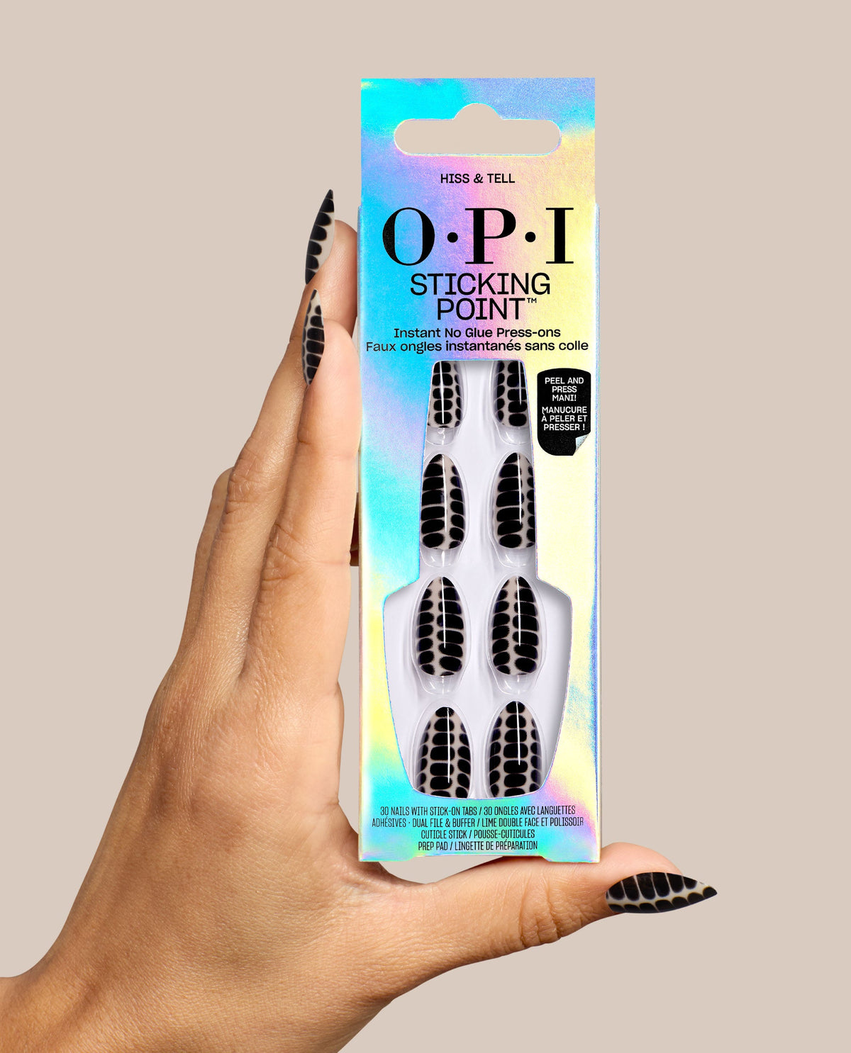 Hand holding OPI Hiss & Tell Sticking Point no-glue press-on nails, featuring black and nude snake-pattern designs in iridescent packaging, styled as bold fall nail colors on a beige background.