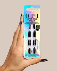 Hand holding OPI Hiss & Tell Sticking Point no-glue press-on nails, featuring black and nude snake-pattern designs in iridescent packaging, styled as bold fall nail colors on a beige background.