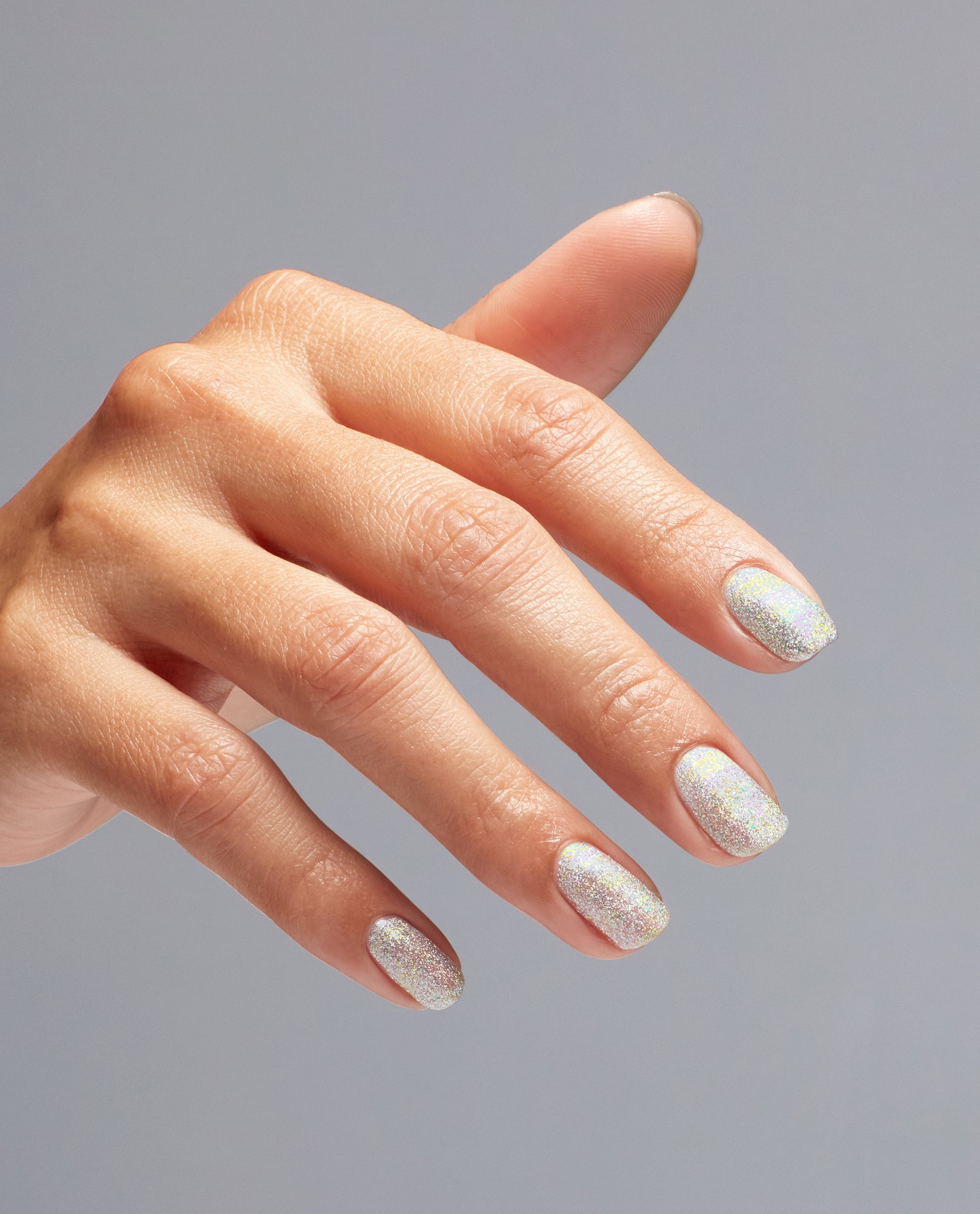 Hand with neatly manicured nails shows OPI nail polish shade I Cancer-tainly Shine, a silver holographic glitter nail lacquer sparkling under soft light against a plain gray background.