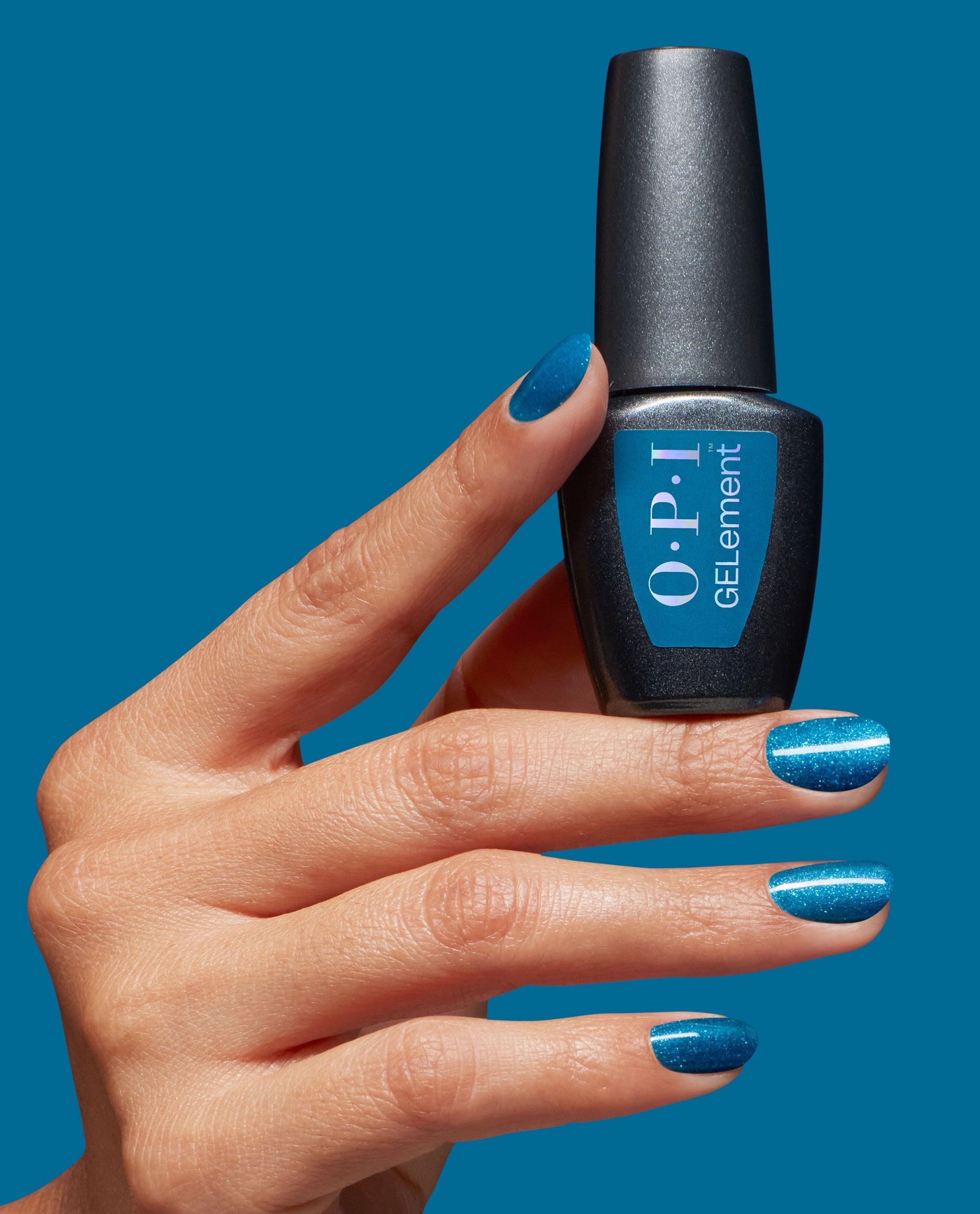 Hand with shimmering teal-blue nails holds an OPI I Sea You at-home gel nail polish bottle against a matching blue background, showcasing a trending ocean-inspired manicure.