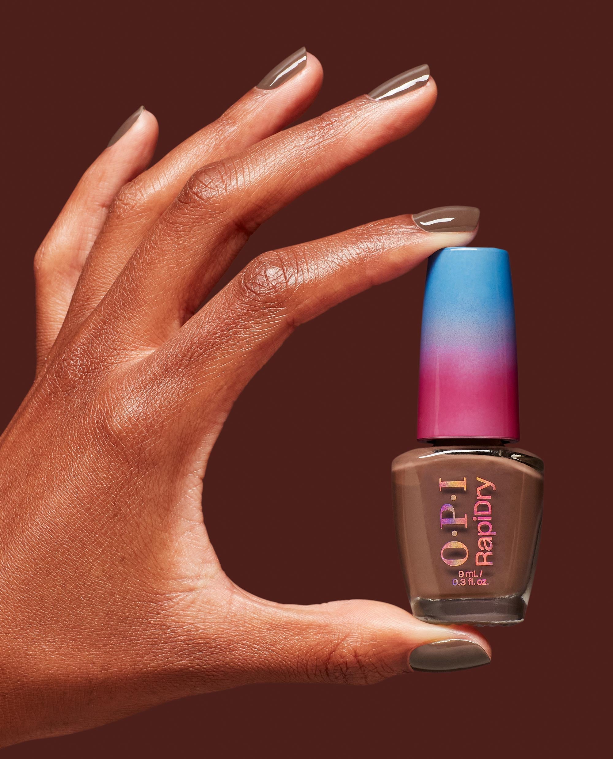 Hand with glossy taupe-brown nails holds an OPI RapiDry bottle in Impromptu You, a quick-dry polish shade for fall nail colors, against a warm brown background.