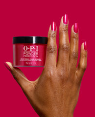 Hand with glossy magenta nails holds OPI Powder Perfection Dipping Powder in It’s All Greek to Me, a vibrant pink shade for bold fall nail colors on a matching pink background.