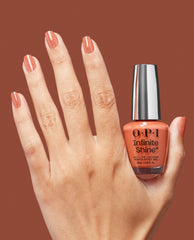 Hand with manicured nails painted in shimmery copper OPI nail polish holds a bottle of OPI Infinite Shine Knee High Stock-blings against a matching warm terracotta background.
