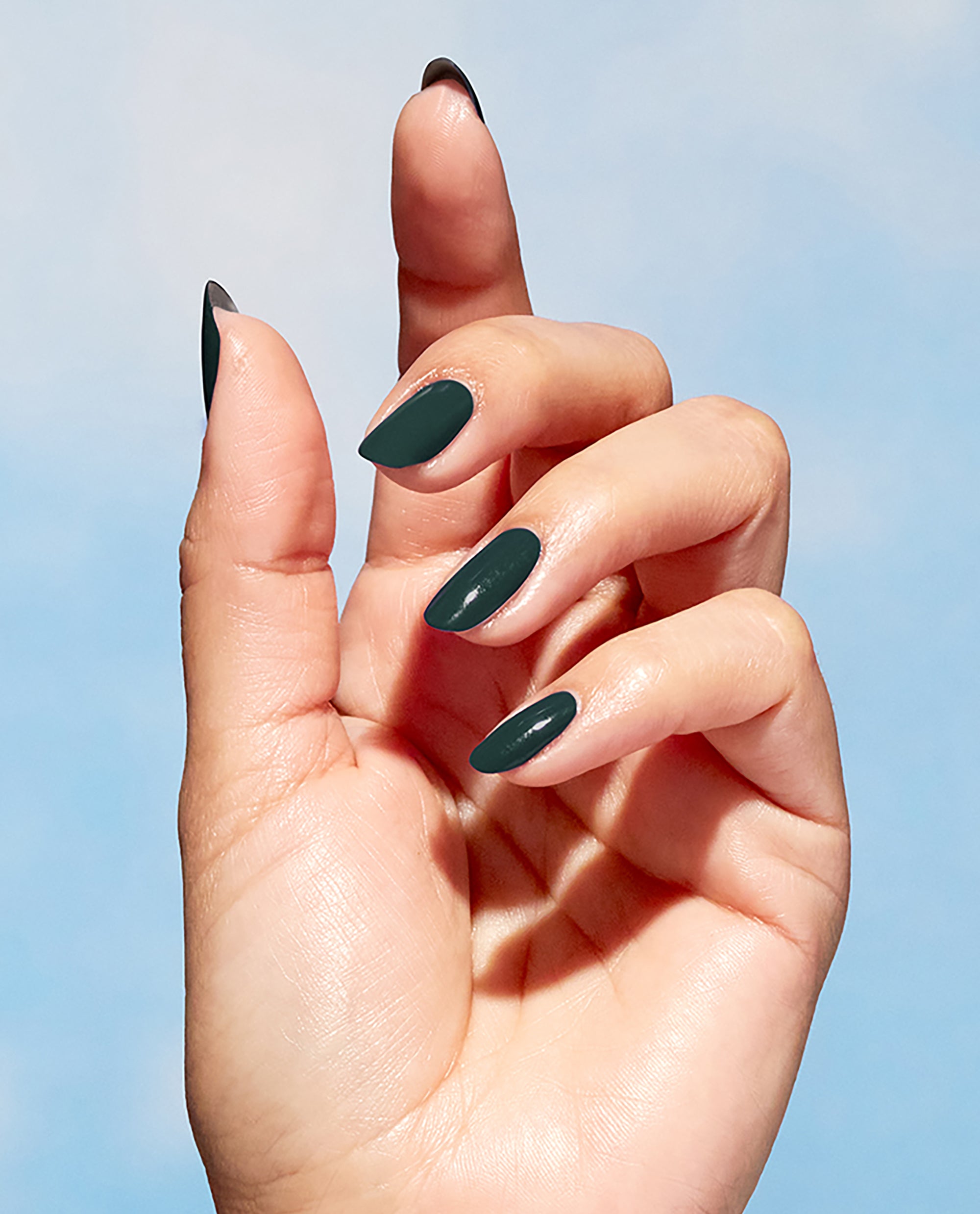 Hand displaying glossy, almond-shaped nails painted a deep forest green, showcasing Leaf by Example Nature Strong Vegan Nail Lacquer by OPI; a minimalist blue-sky backdrop emphasizes the rich green nail polish, perfect for spring nail colors.