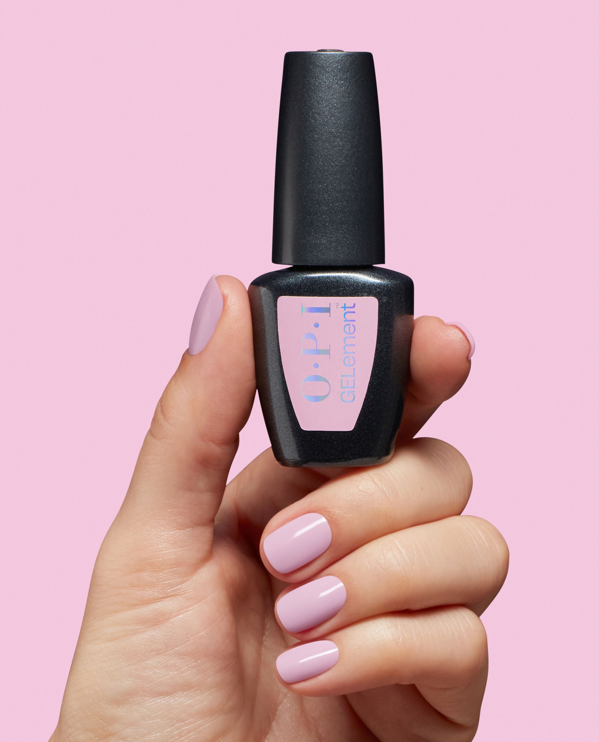 Hand with glossy lavender manicure holds OPI Let Me Lavender at-home gel nail polish bottle against a pink background, showcasing a soft shade for fall nail colors.