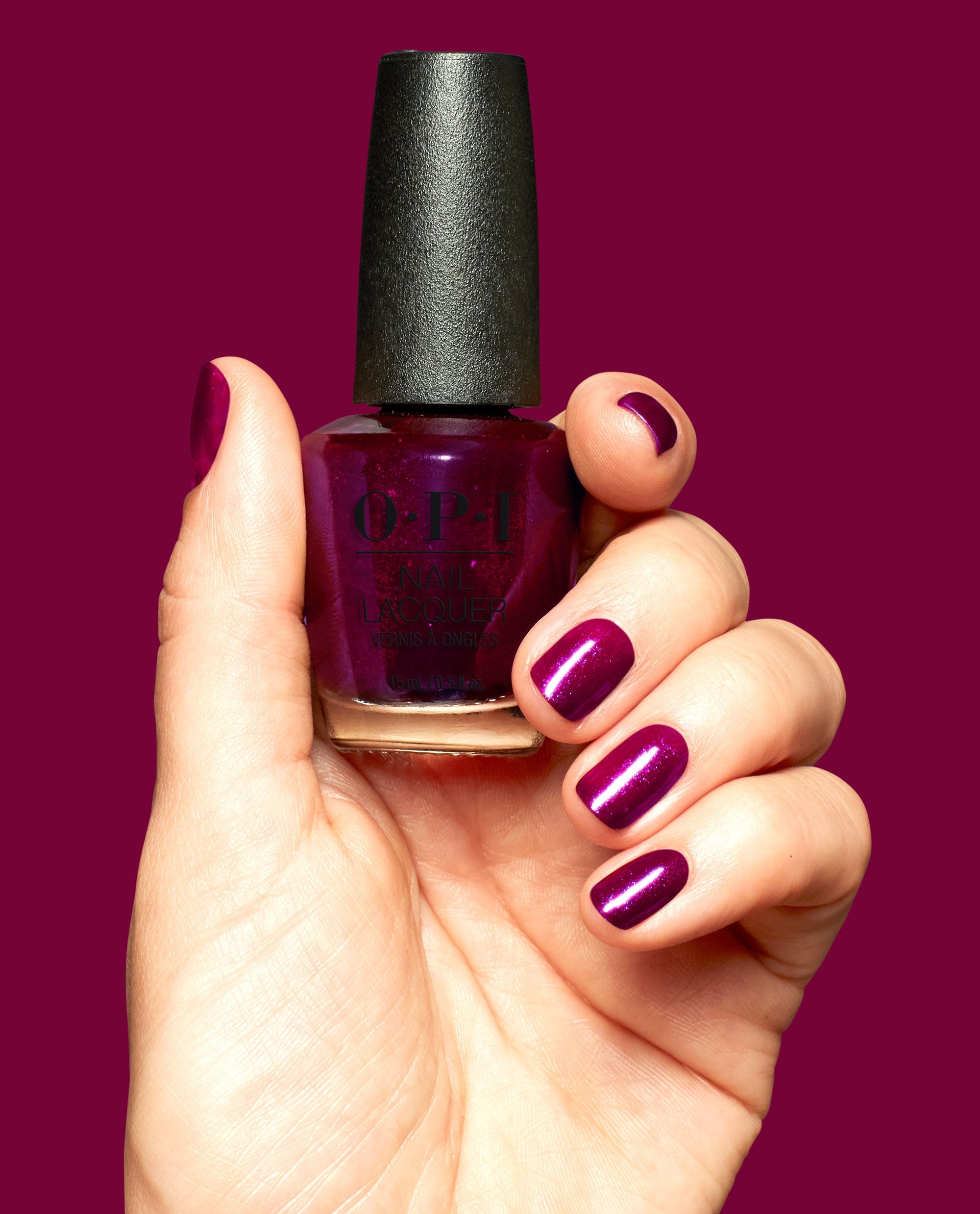 Hand with glossy deep berry-purple nails holds an OPI Lincoln Park After Brunch Nail Lacquer bottle, showing matching shimmer polish against a solid burgundy background.