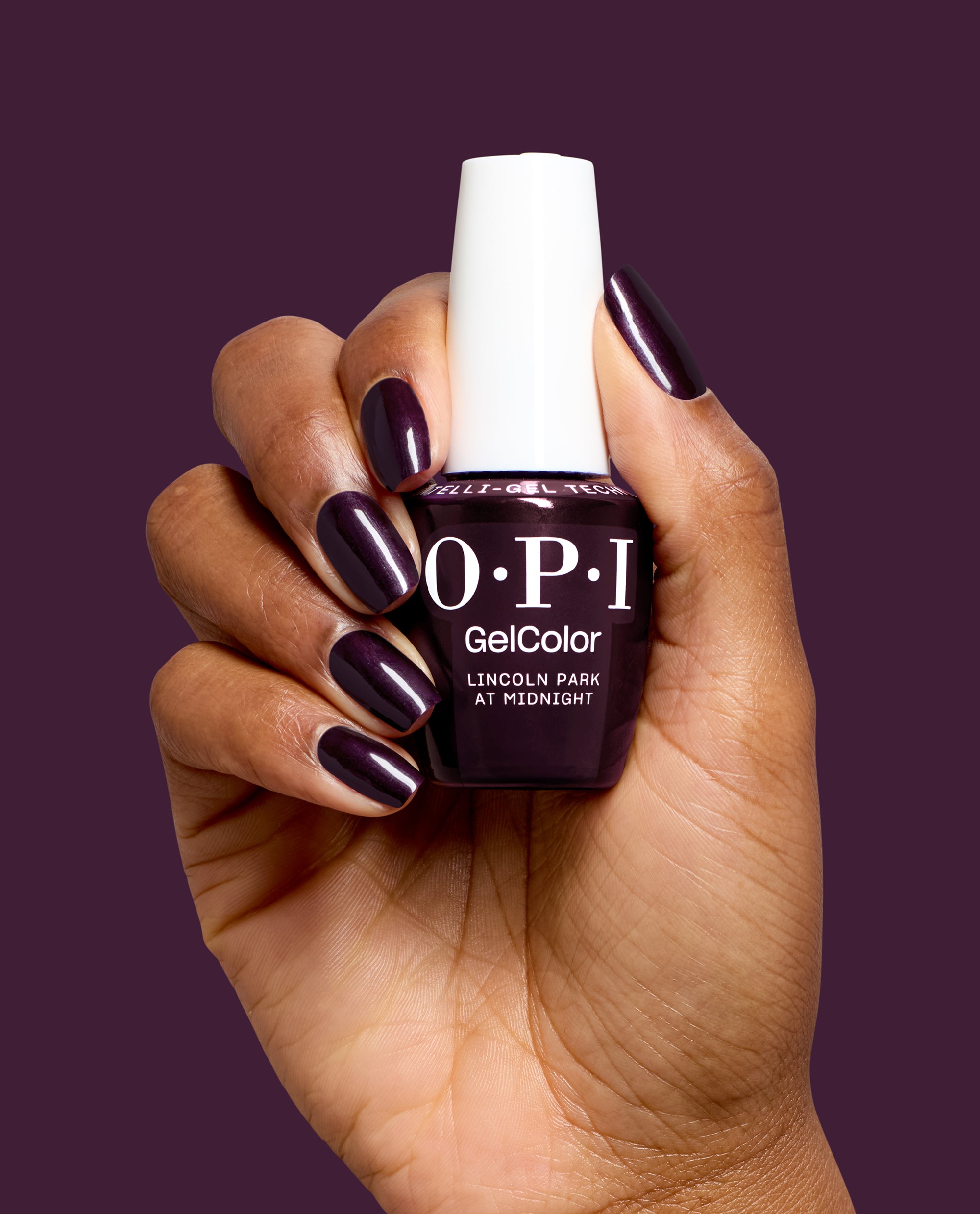 Hand with glossy deep plum nails holds OPI GelColor Lincoln Park at Midnight gel nail polish, a dark purple shade for chic fall nail colors, against a matching plum background.