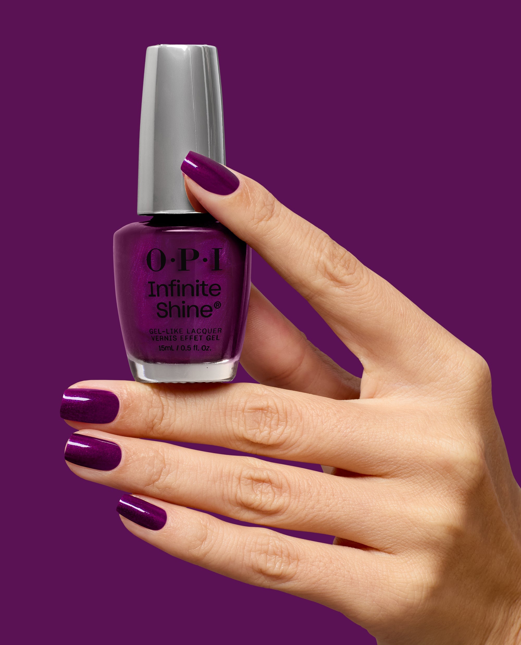 Hand with glossy deep plum nails holds an OPI Infinite Shine bottle in Louvre Me Lourve Me Not, a rich purple gel-like lacquer shade for fall nail colors, against a matching purple background.