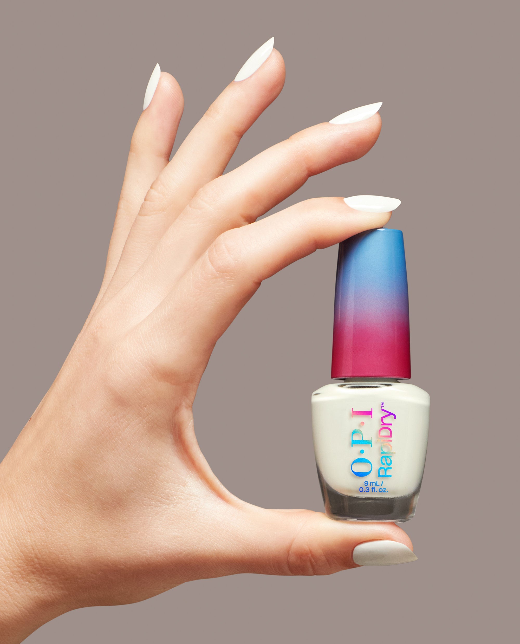 Manicured hand with white nails holding an OPI RapiDry Love at Frost Sight nail polish bottle with a pink-to-blue ombré cap against a neutral background, showcasing a chic OPI nail polish gift set for fast-drying shine.