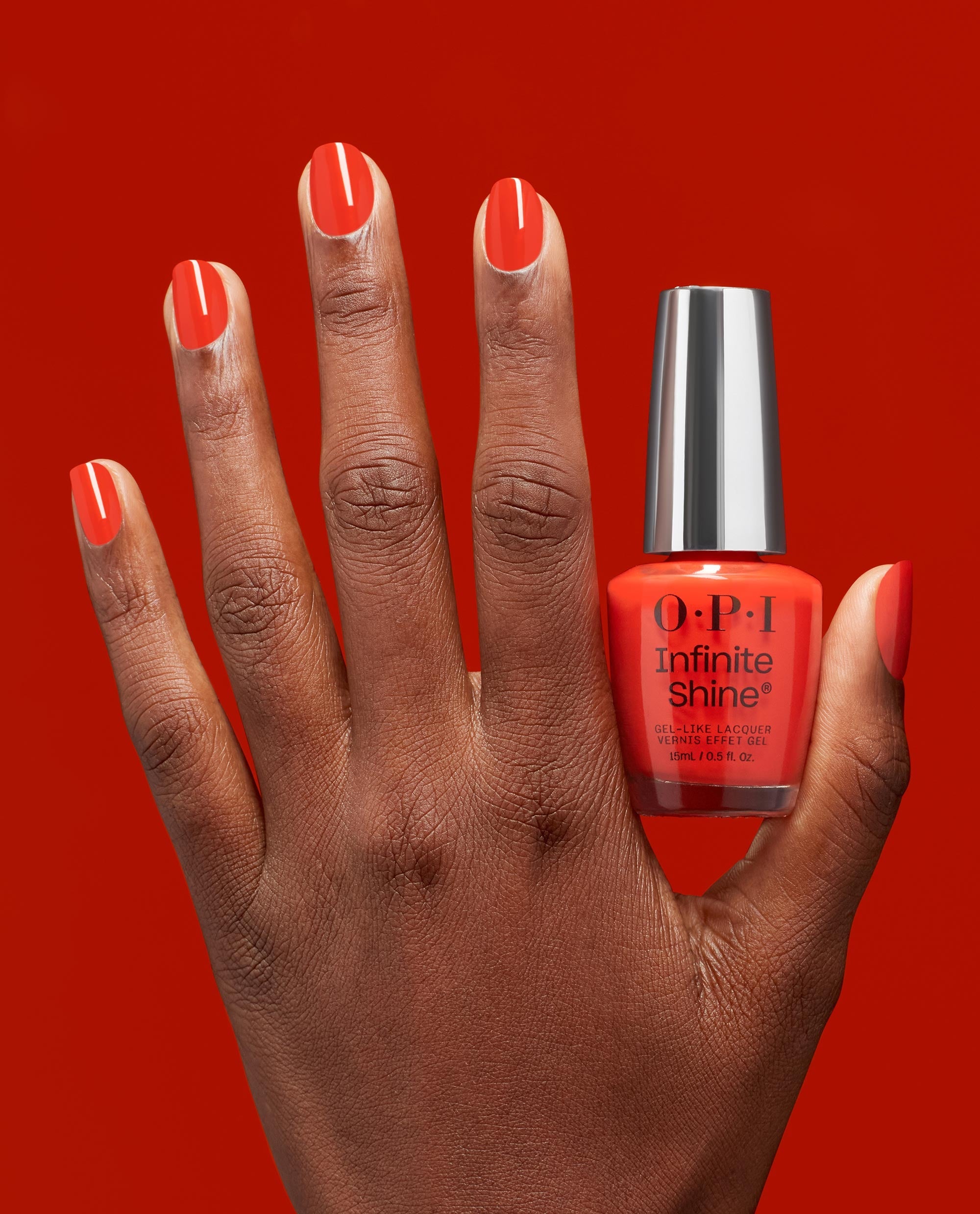 Hand with glossy coral-red nails holds a Mini Plaid Flirt Infinite Shine bottle of OPI nail polish against a matching bold red background, showcasing the vibrant gel-like lacquer color and shine.