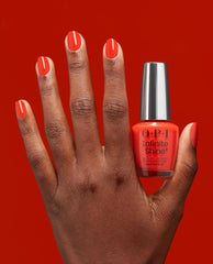 Hand with glossy coral-red nails holds a Mini Plaid Flirt Infinite Shine bottle of OPI nail polish against a matching bold red background, showcasing the vibrant gel-like lacquer color and shine.