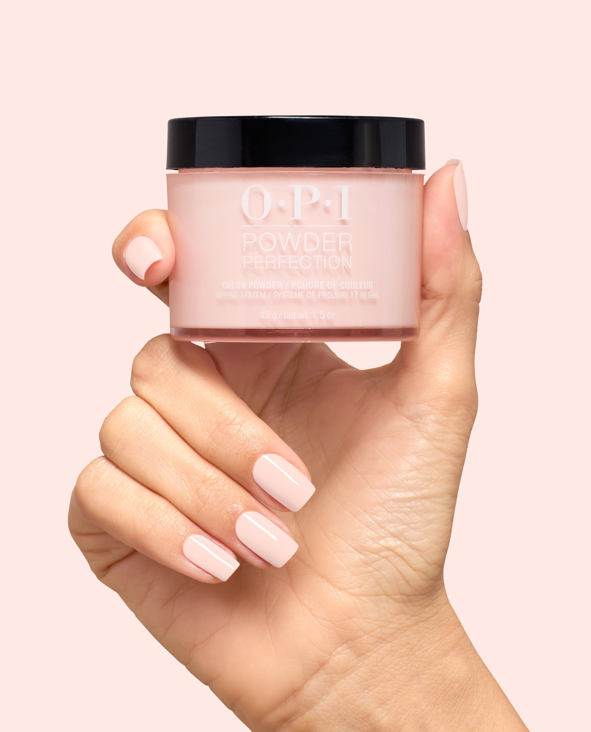 Hand with pale pink manicured nails holds OPI Powder Perfection Mod About You Pastel dipping powder jar, a soft blush shade for OPI fall nail colors, on a light pink background.