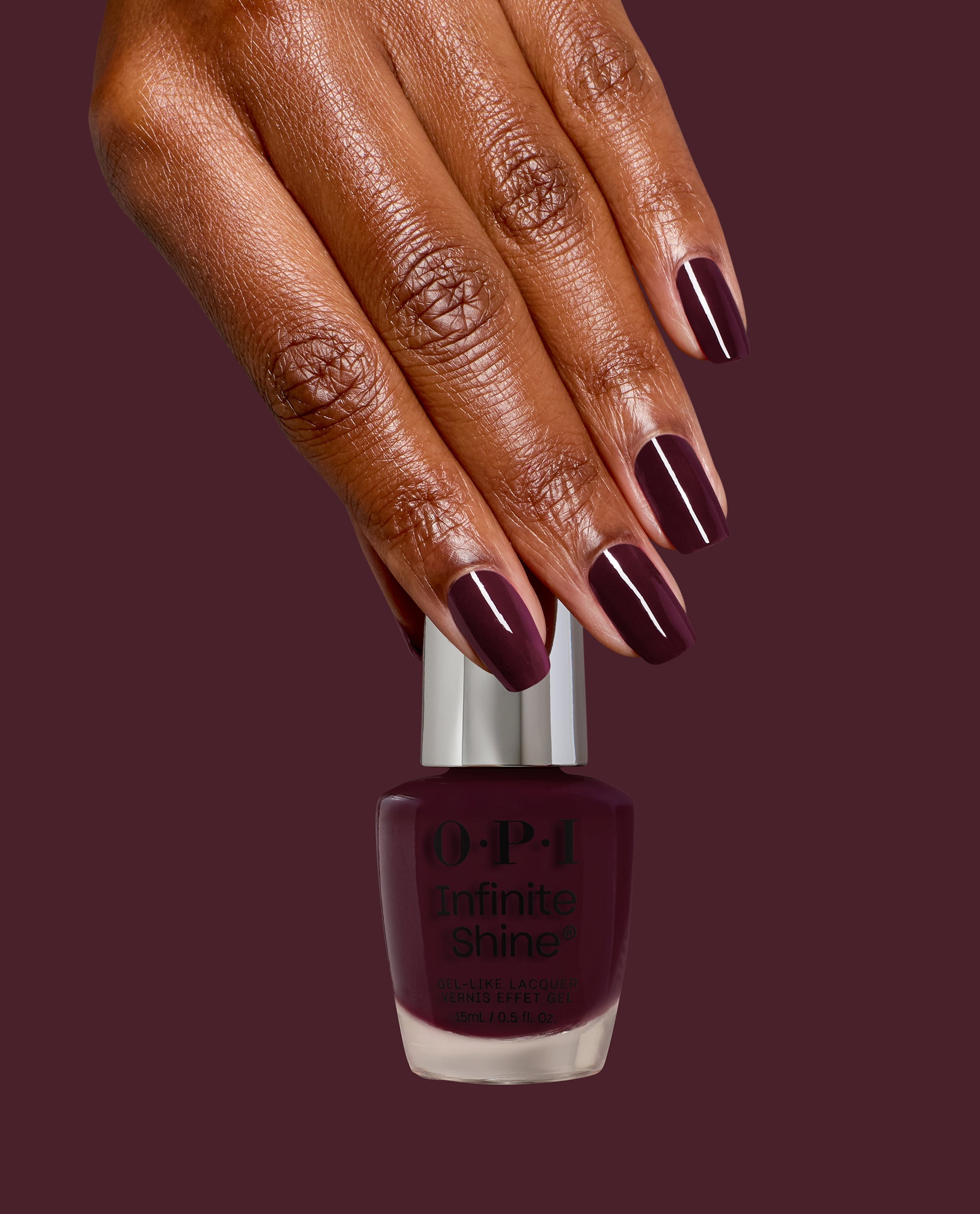 Hand with glossy deep burgundy nails holds OPI Infinite Shine Mrs. O'Leary's BBQ nail polish bottle, showcasing rich fall nail colors on a matching wine background.