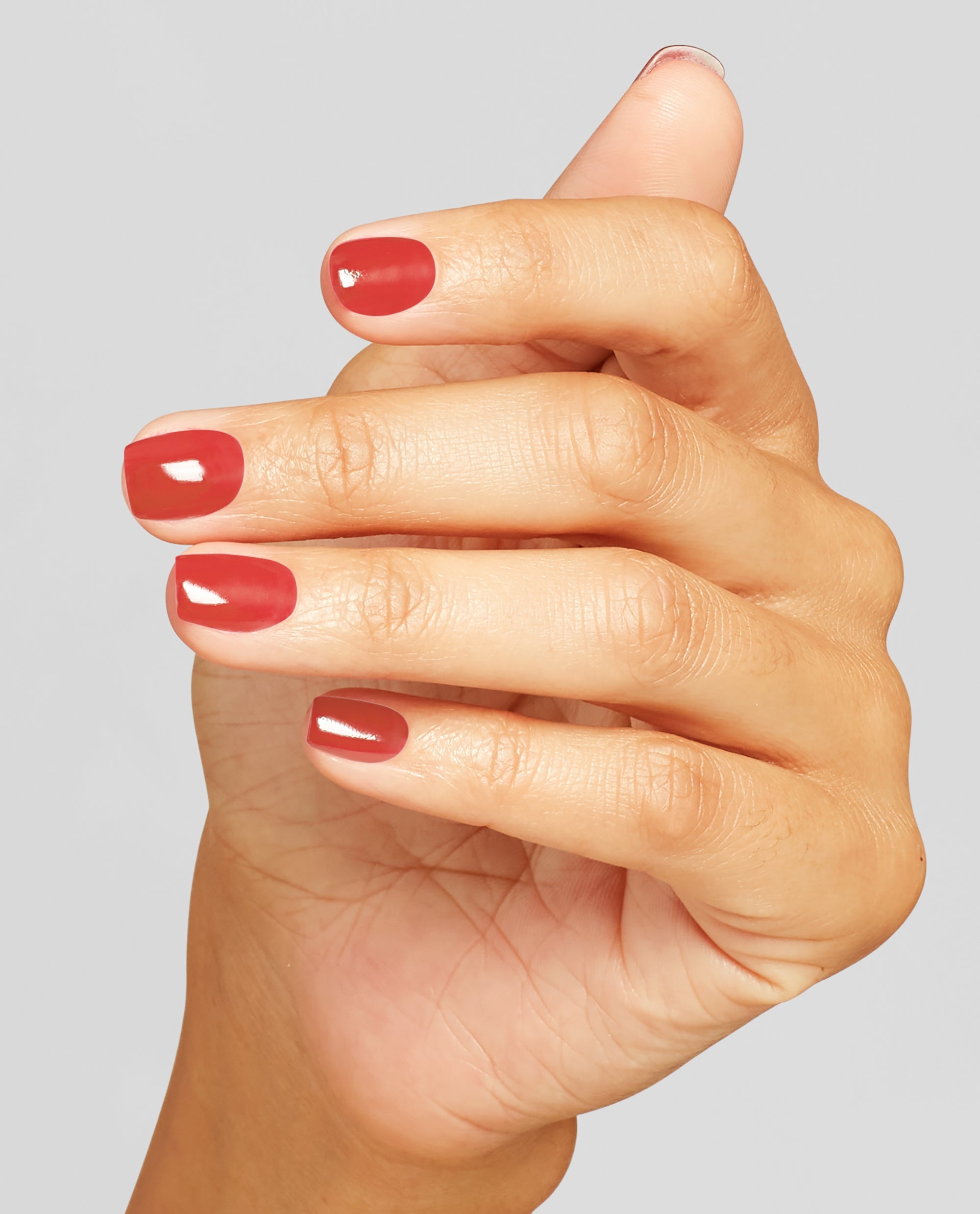 Hand with short, neatly manicured nails painted in OPI nail polish shade My Solar Clock is Ticking, a warm terracotta-red nail lacquer, held against a light gray background to showcase the glossy color and smooth finish.