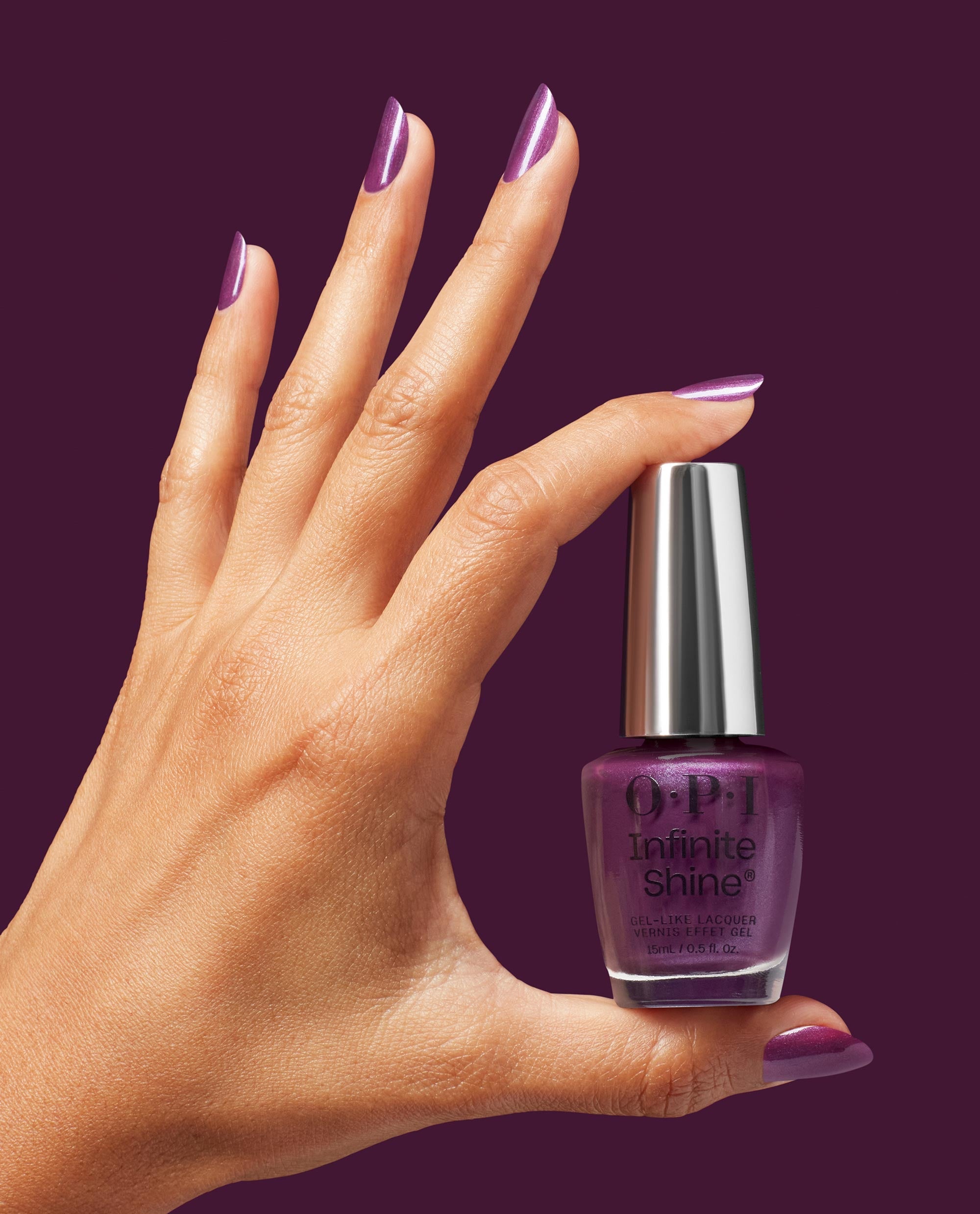 Hand with glossy purple berry nails holds OPI Infinite Shine My Berry Janes gel-like lacquer bottle against a deep plum background, showcasing rich fall nail colors.