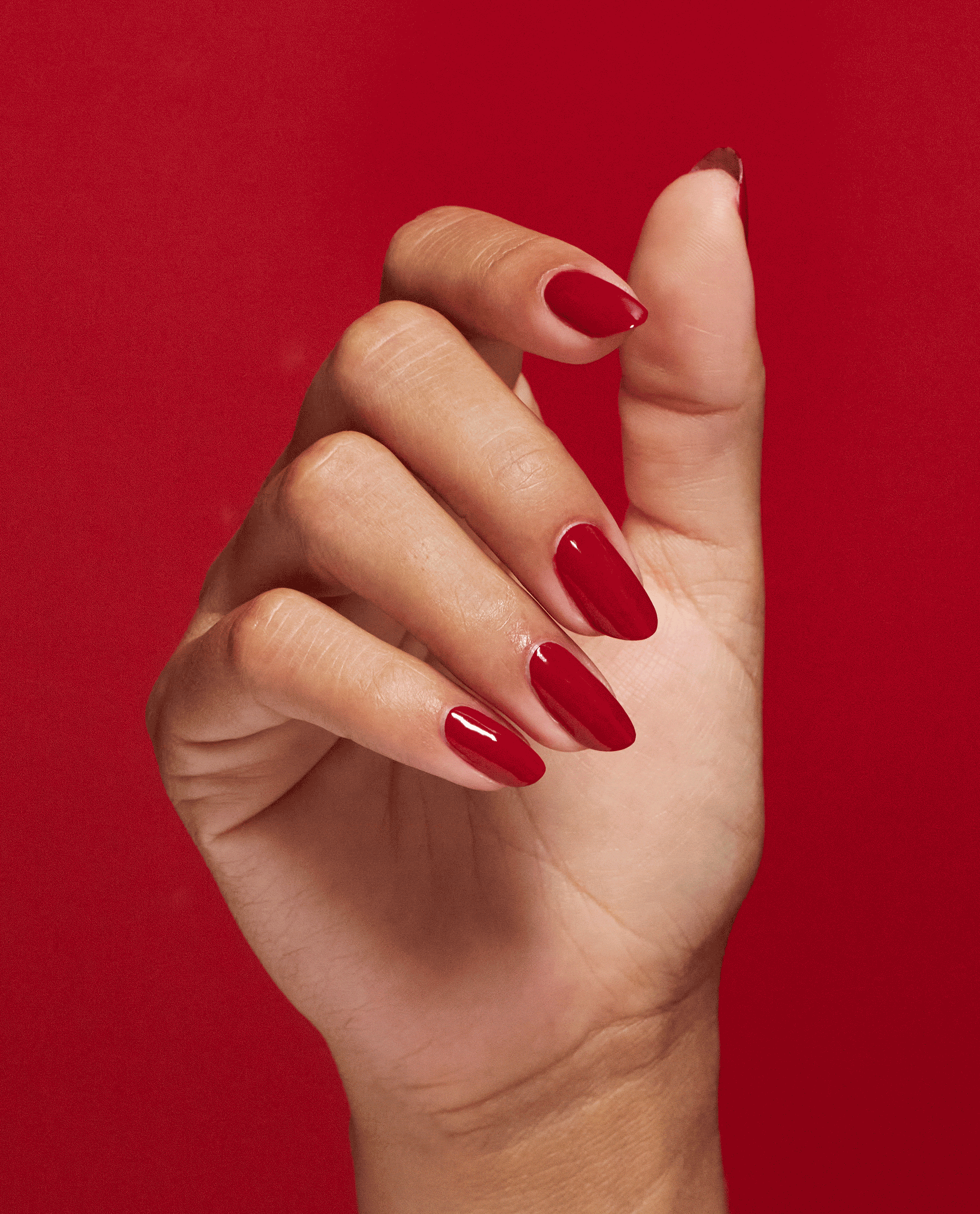 Hand displays glossy almond-shaped red nails with OPI Nail Envy Big Apple Red nail strengthener, a bold treatment shade suited for fall nail colors, against a rich red background.