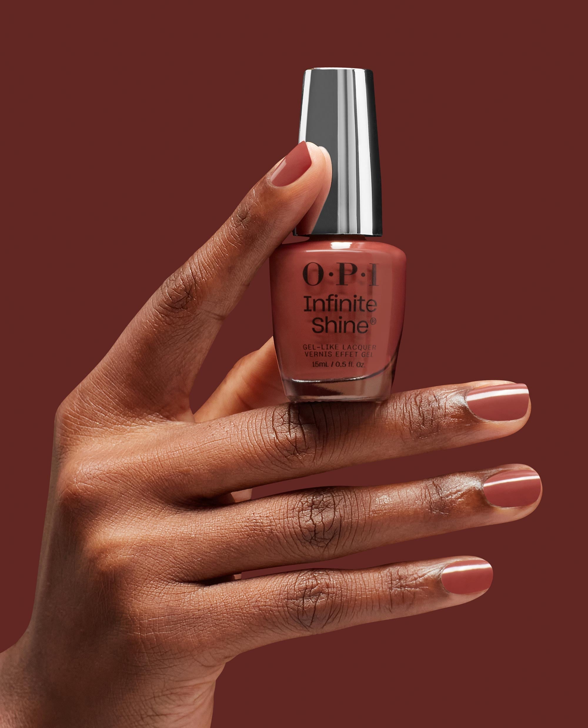OPI Infinite Shine Nail Prep Rally gel-like lacquer bottle held by a hand with glossy warm terracotta nails, showcasing a rich shade for fall nail colors on a matching brown background.