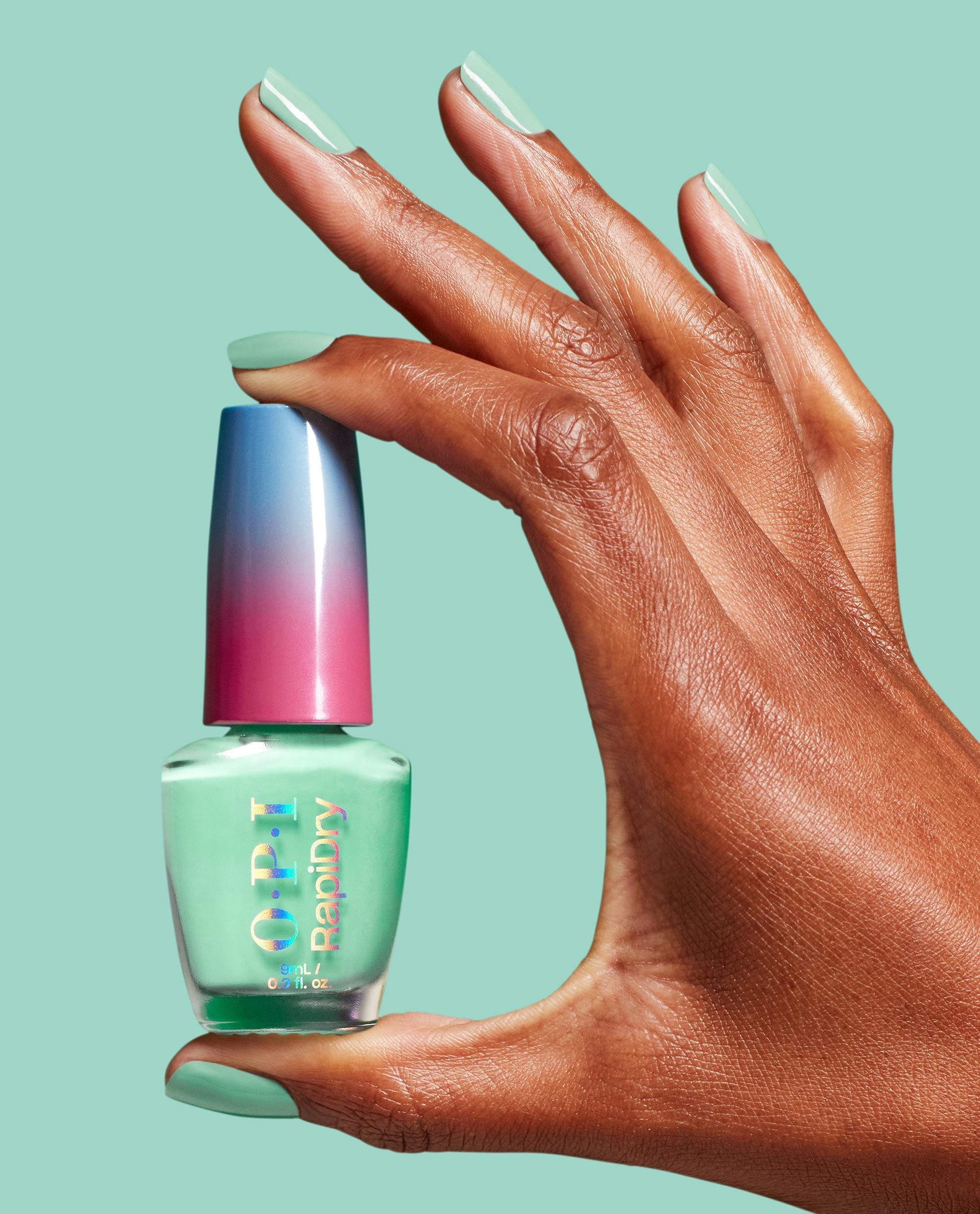OPI RapiDry™ No Further Delays green nail polish bottle held between a manicured hand’s fingers, nails painted mint green, against a matching mint background with a blue-to-pink gradient cap, showcasing fresh spring nail colors by OPI.
