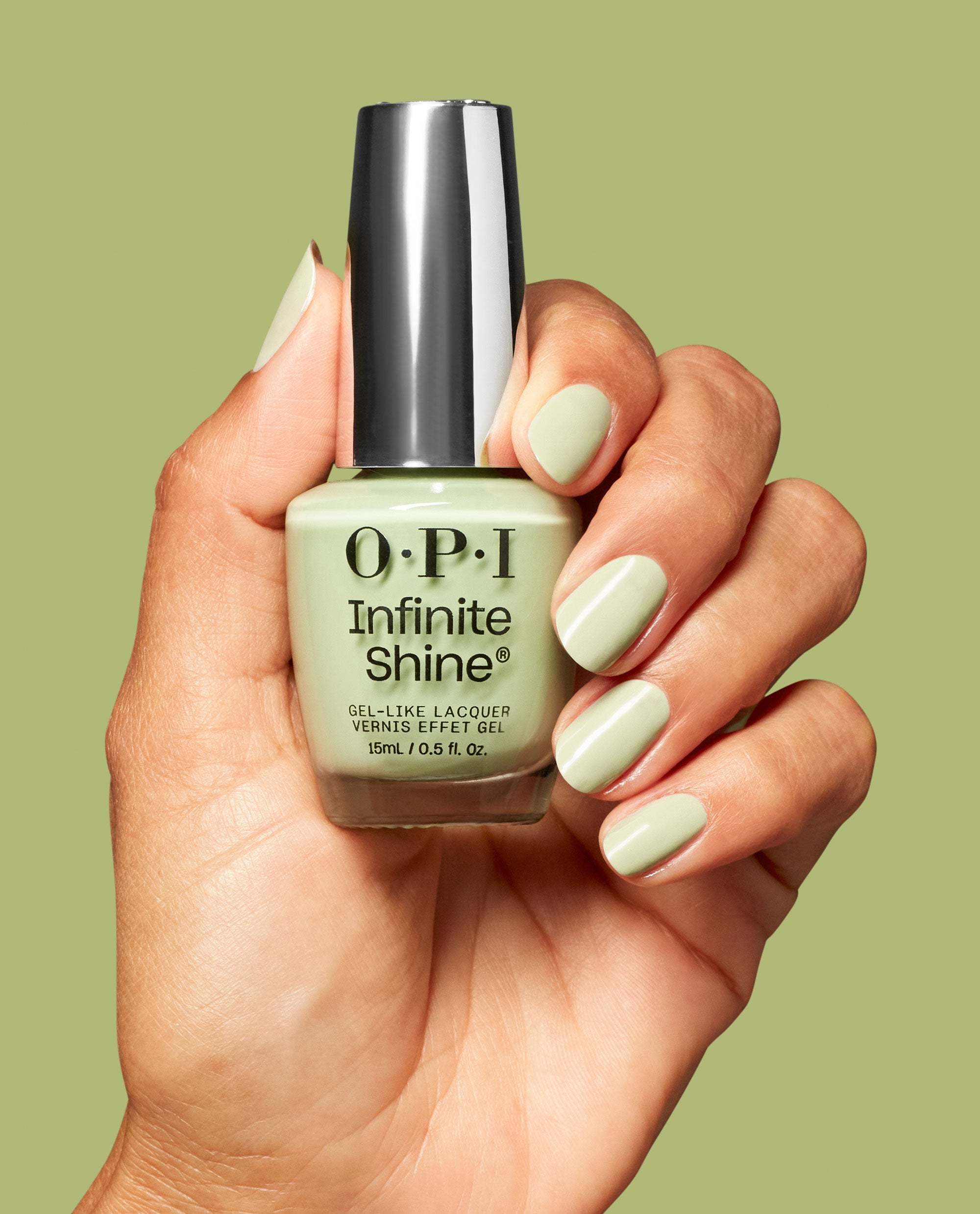 Hand with glossy pale green nails holds OPI Infinite Shine Oh Baby! gel-like lacquer bottle against a matching green background.