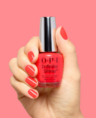 Hand with glossy coral-red nails holds OPI Infinite Shine Pack a Cherry-on Bag gel-like lacquer bottle against a pink background, showcasing a trending nail polish shade.