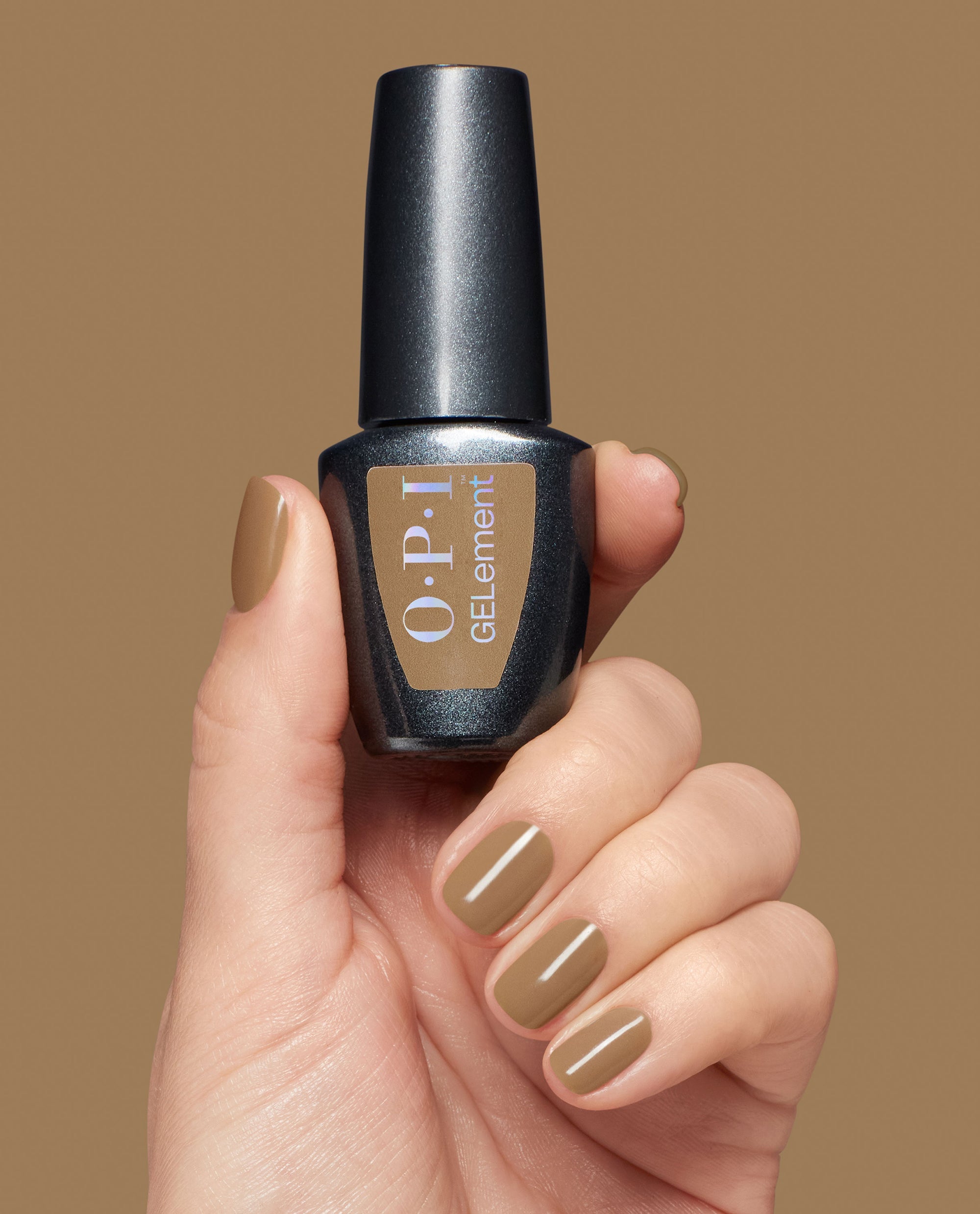 Hand with glossy caramel-brown nails holds OPI GelColor Peanut Butter & Gel-ly at-home gel nail polish bottle against a warm tan background, showcasing cozy fall nail colors.