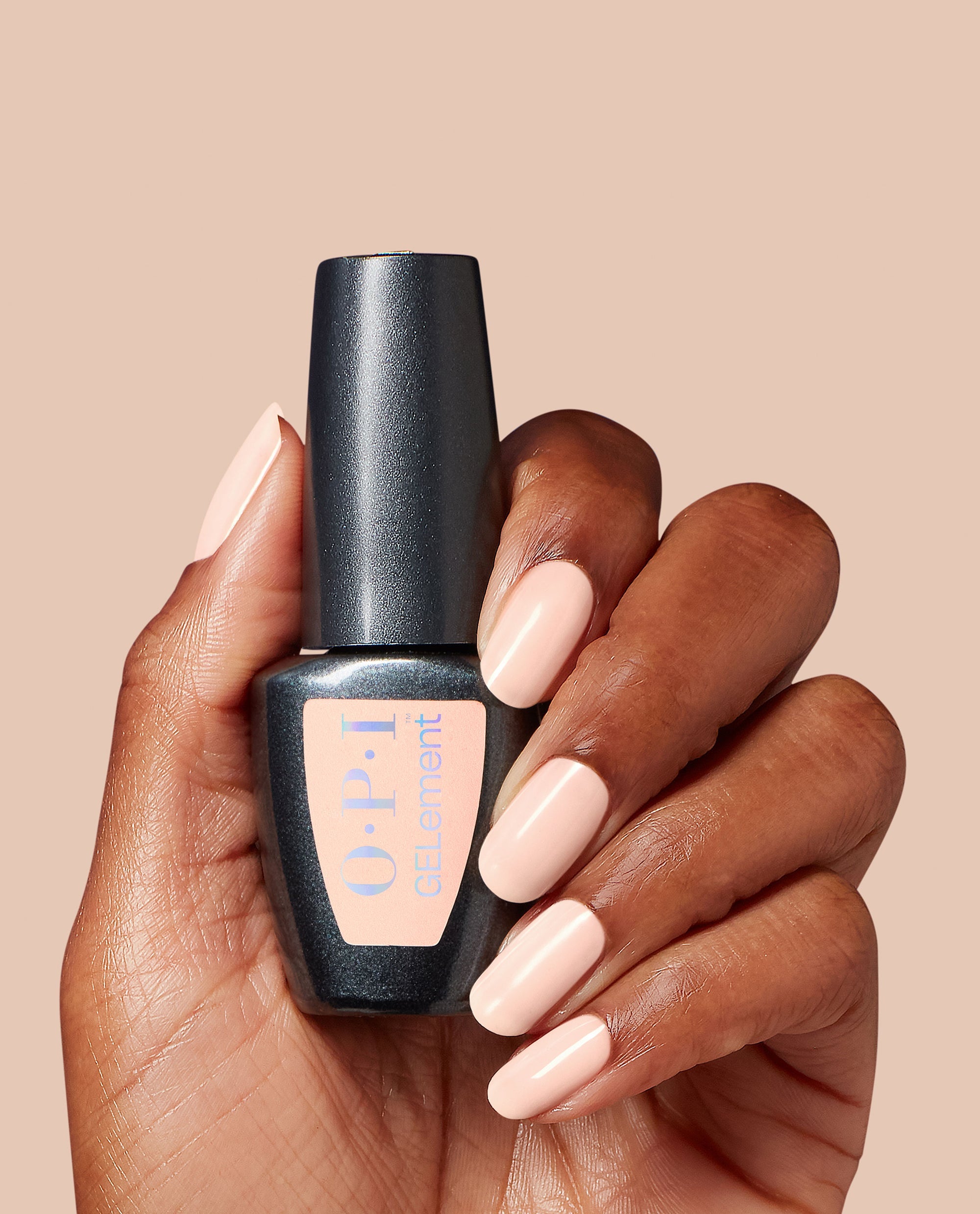 Hand with glossy pale pink Pilates Princess at-home gel manicure holding an OPI GelElement bottle against a nude background, showcasing the soft neutral shade as part of an elegant OPI nail polish gift set for salon-quality nails at home.